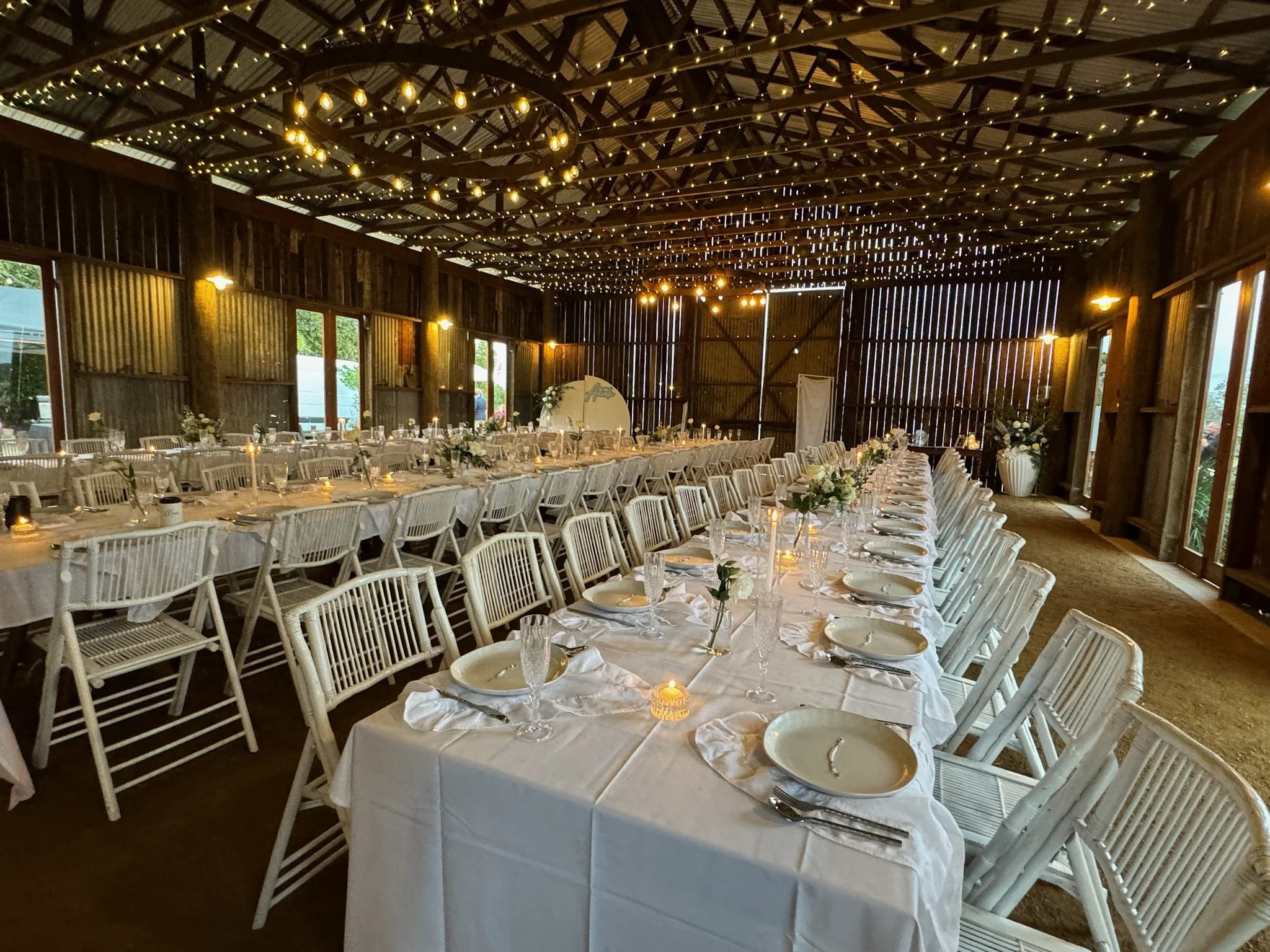 Long rectangular banquet table set for a wedding reception in a rustic barn with fairy lights and floral centerpieces.