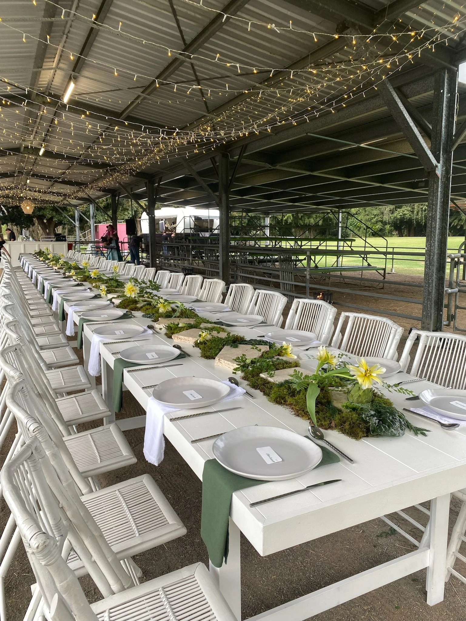 Long rectangular dining table set with white plates, silverware, green napkins, and white chairs in a covered outdoor venue decorated with string lights and green foliage.