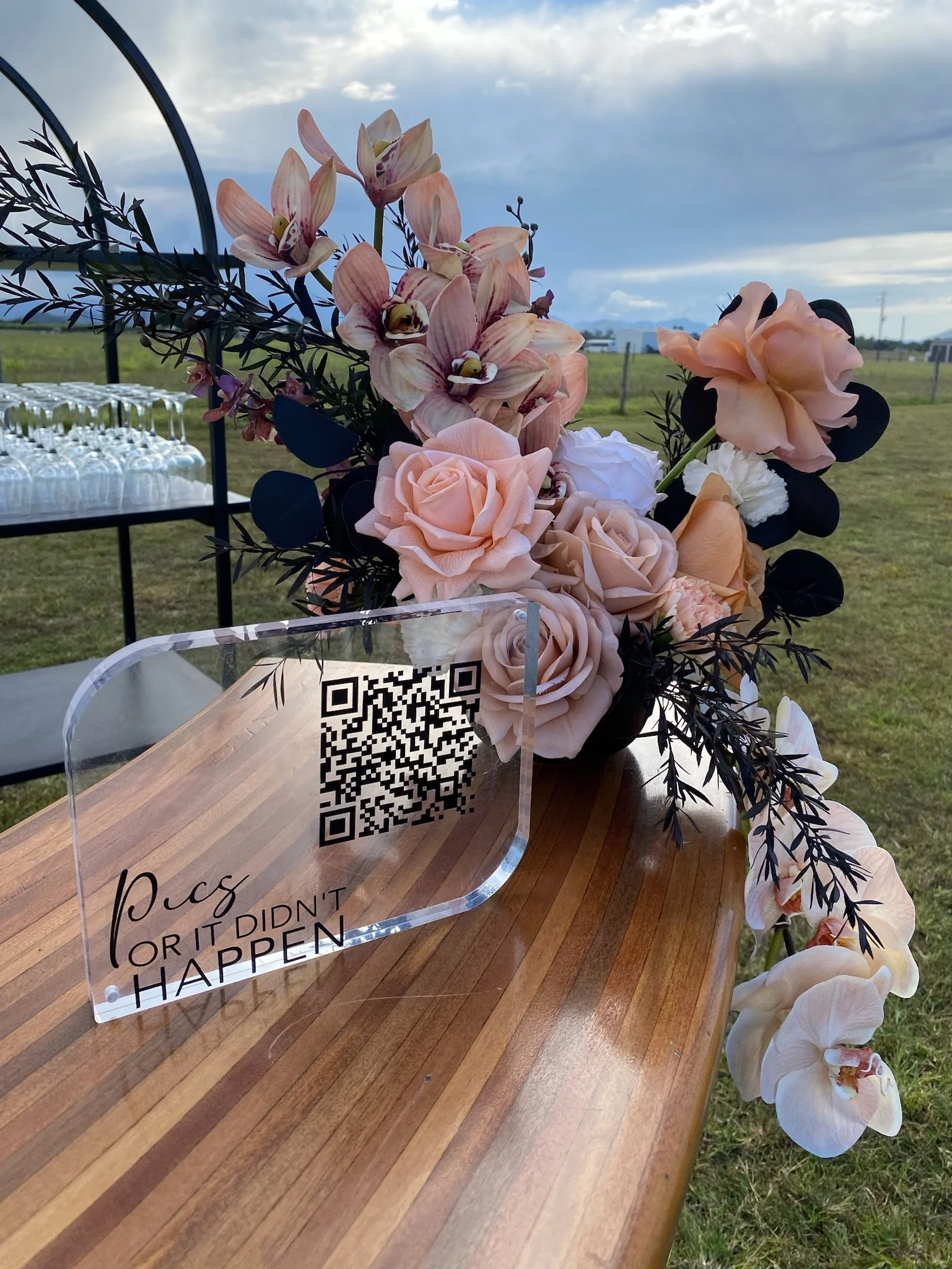 A floral arrangement of peach, white, and pink roses, orchids, and foliage on a wooden table outdoors with a sign displaying a QR code and the words 'P.s. (or) it didn't happen' against a background of open green fields and a cloudy sky.