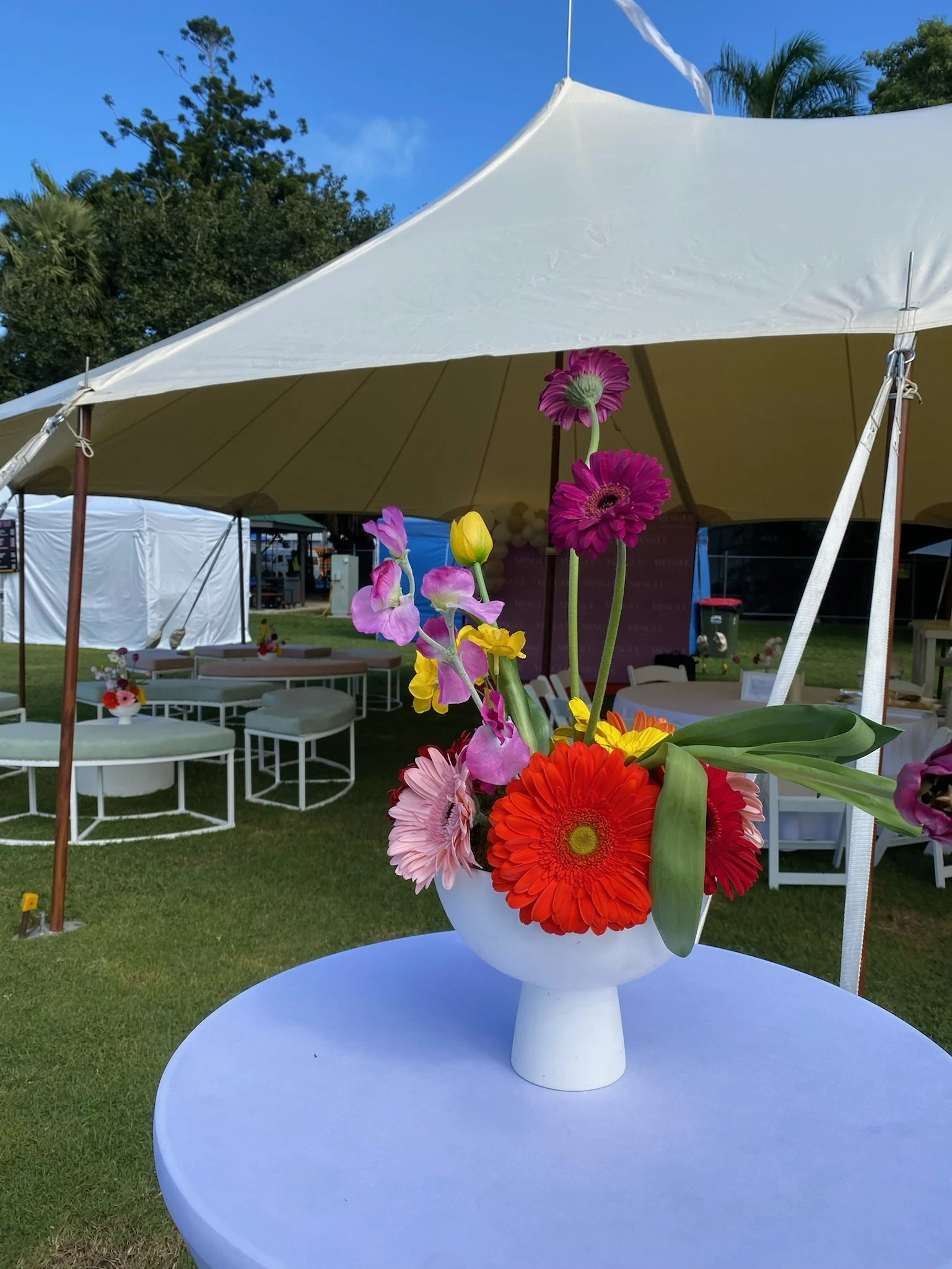 Colorful flower arrangement in a white vase on a table outdoors under a large white tent with additional tables and chairs in the background, greenery, and blue sky.