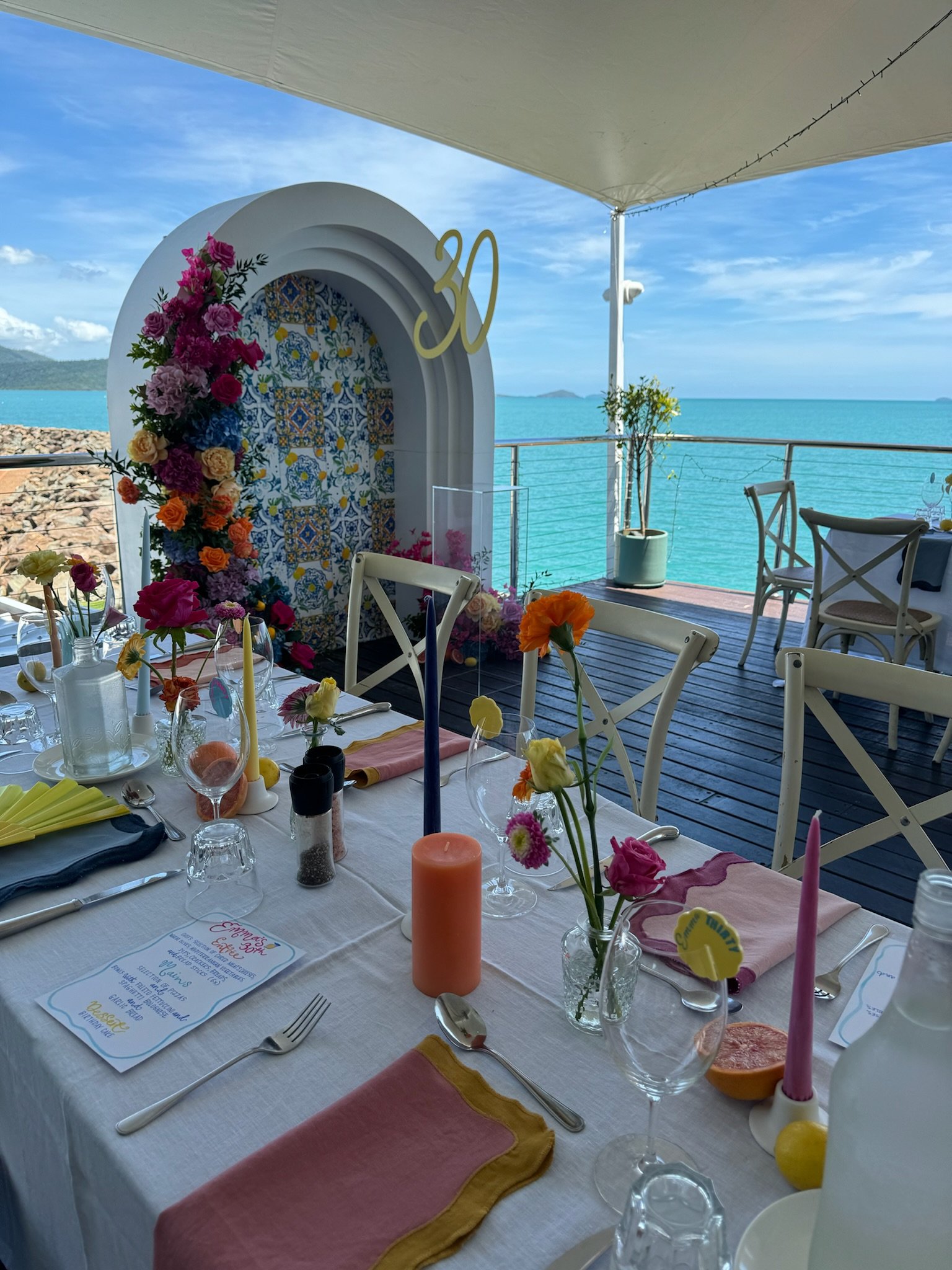Decorated table set for a celebration with flowers, candles, and a menu, overlooking the ocean with blue sky and distant islands.