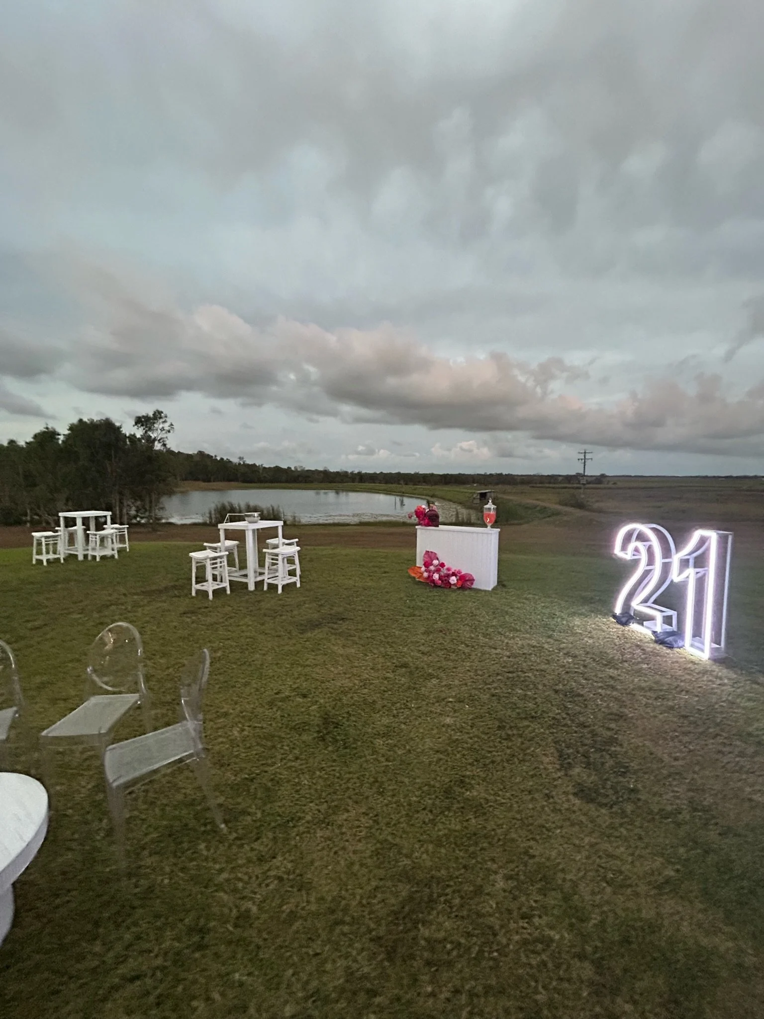 Outdoor wedding or event setup on a grassy area with white tables and chairs, a water body in the background, large illuminated '21' sign, and floral arrangements, under a cloudy sky.