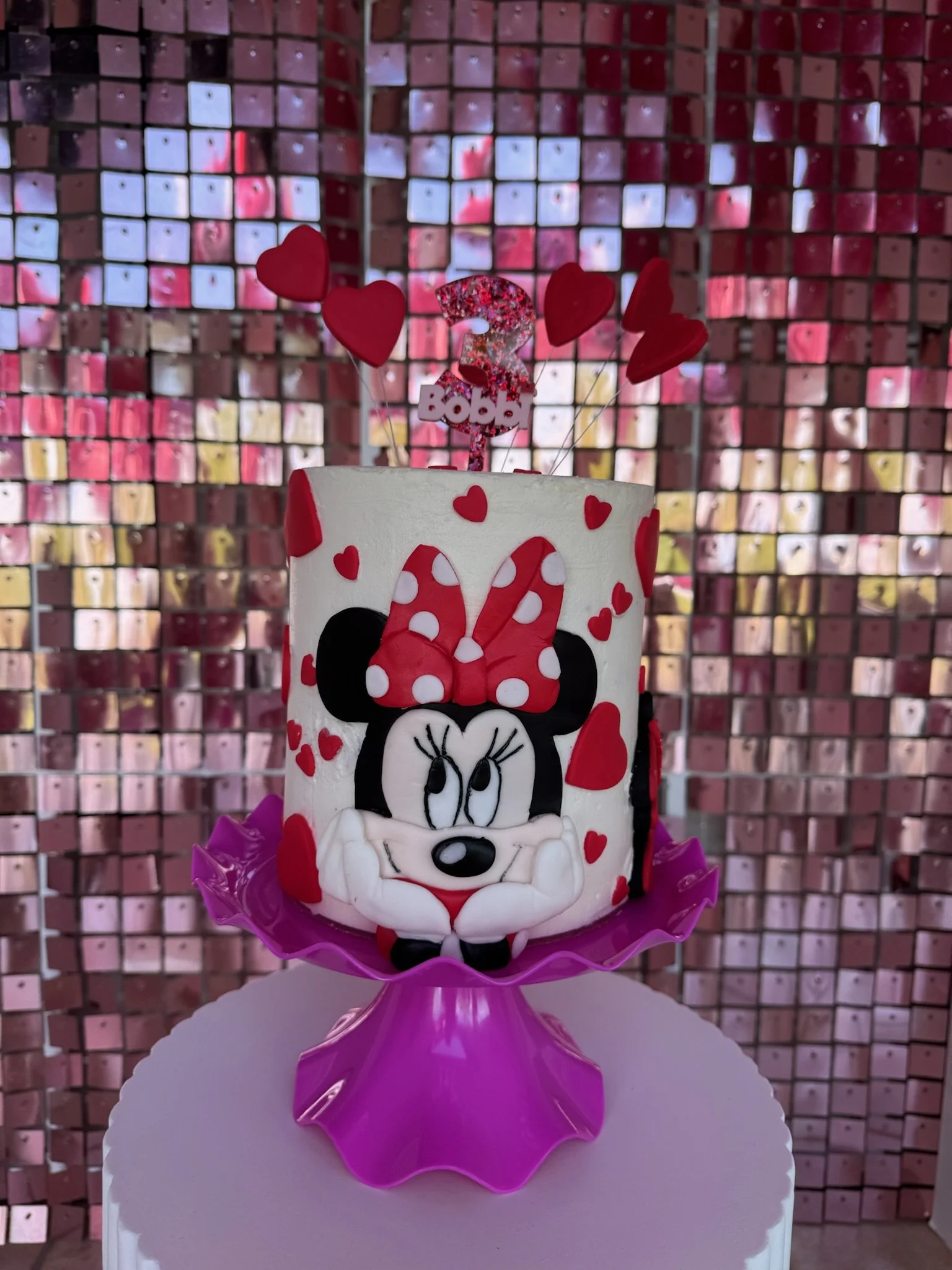 A birthday cake decorated with Minnie Mouse theme, featuring a Minnie face with a red bow with white polka dots, red and pink heart decorations, and a topper with a glitter number 2, red hearts, and a "Baba" sign. The cake sits on a pink plastic base