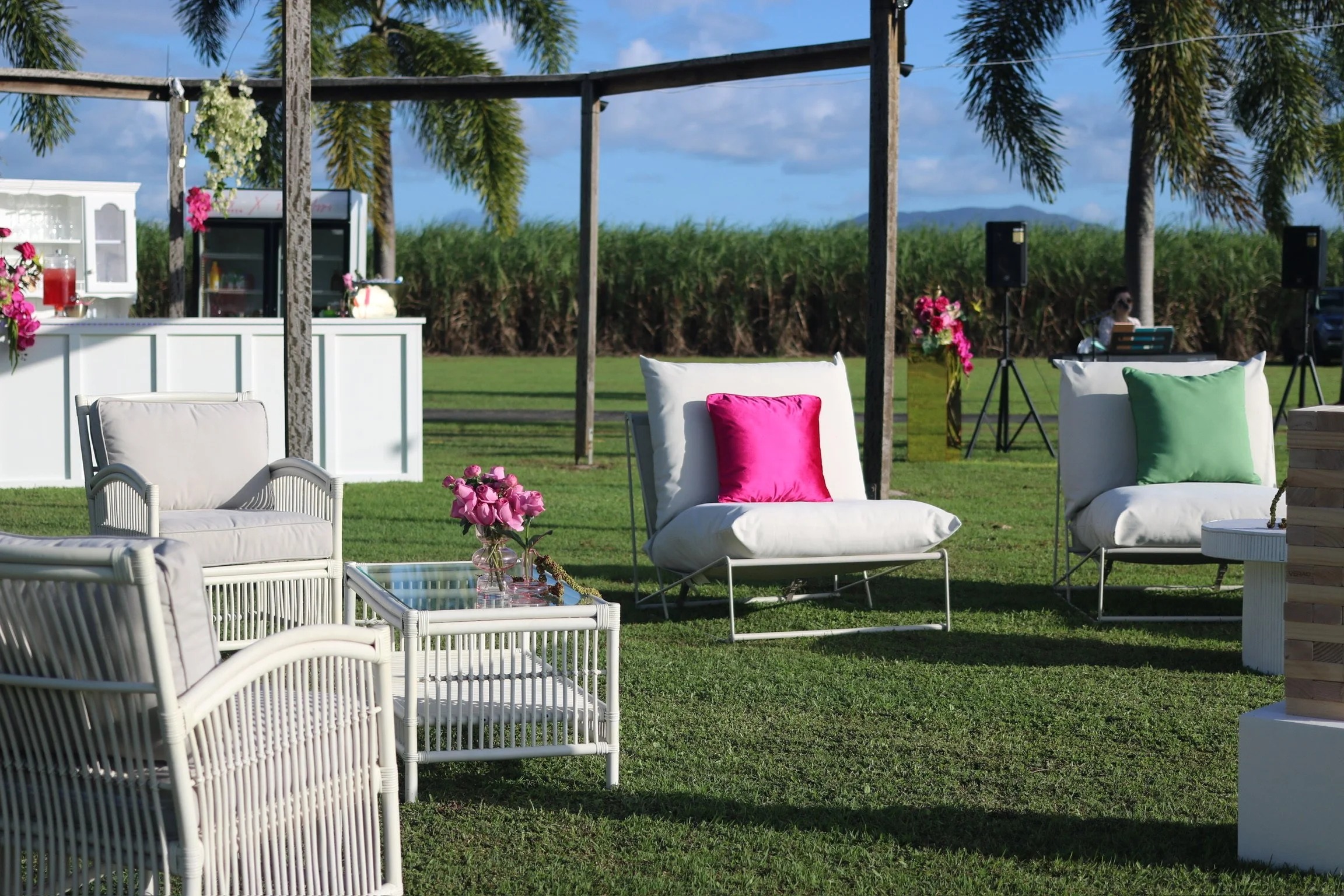 Outdoor event setup with white cushioned chairs and sofas, pink and green throw pillows, a small glass table with a pink flower arrangement, a white bar area, palm trees, and a stage with speakers, set in a grassy field with mountains in the backgrou