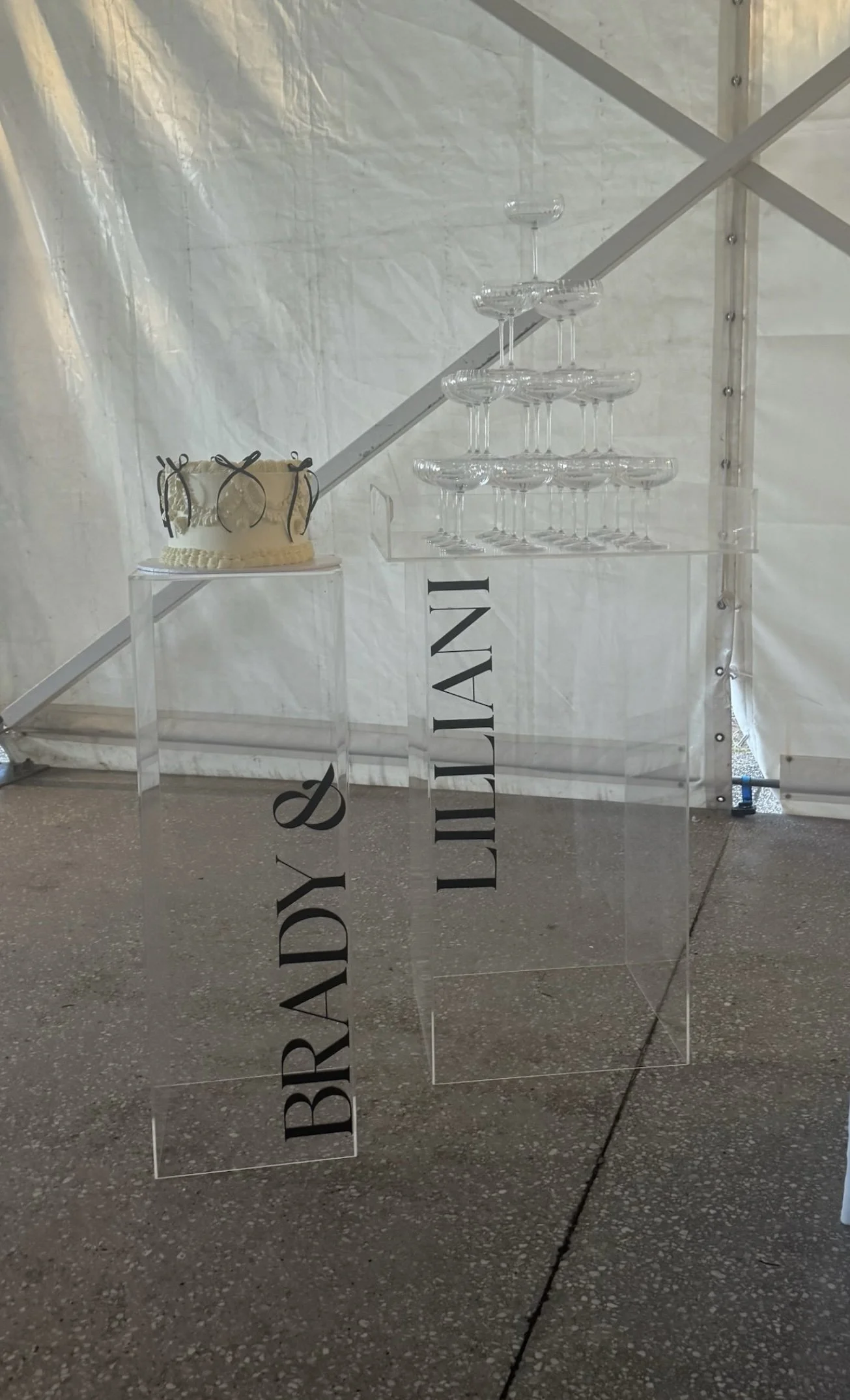 A transparent acrylic stand displays a cake, champagne glasses, and a sign reading 'BRADY & DILLON' at an event wedding or celebration.