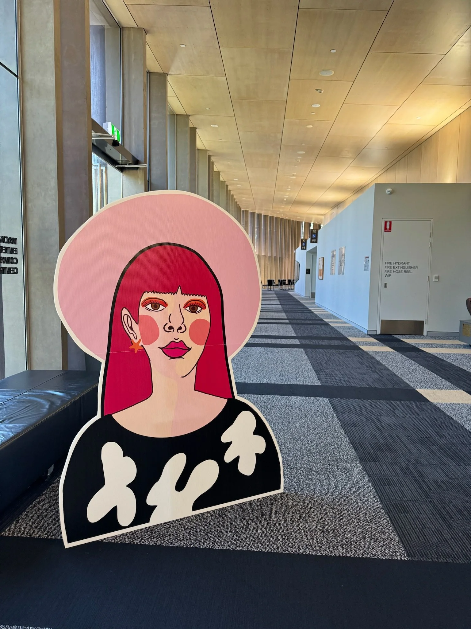 Colorful illustration of a woman with pink hair, wearing a wide pink hat, earrings, and a black blouse with white patterns, placed in an indoor space with modern architecture.