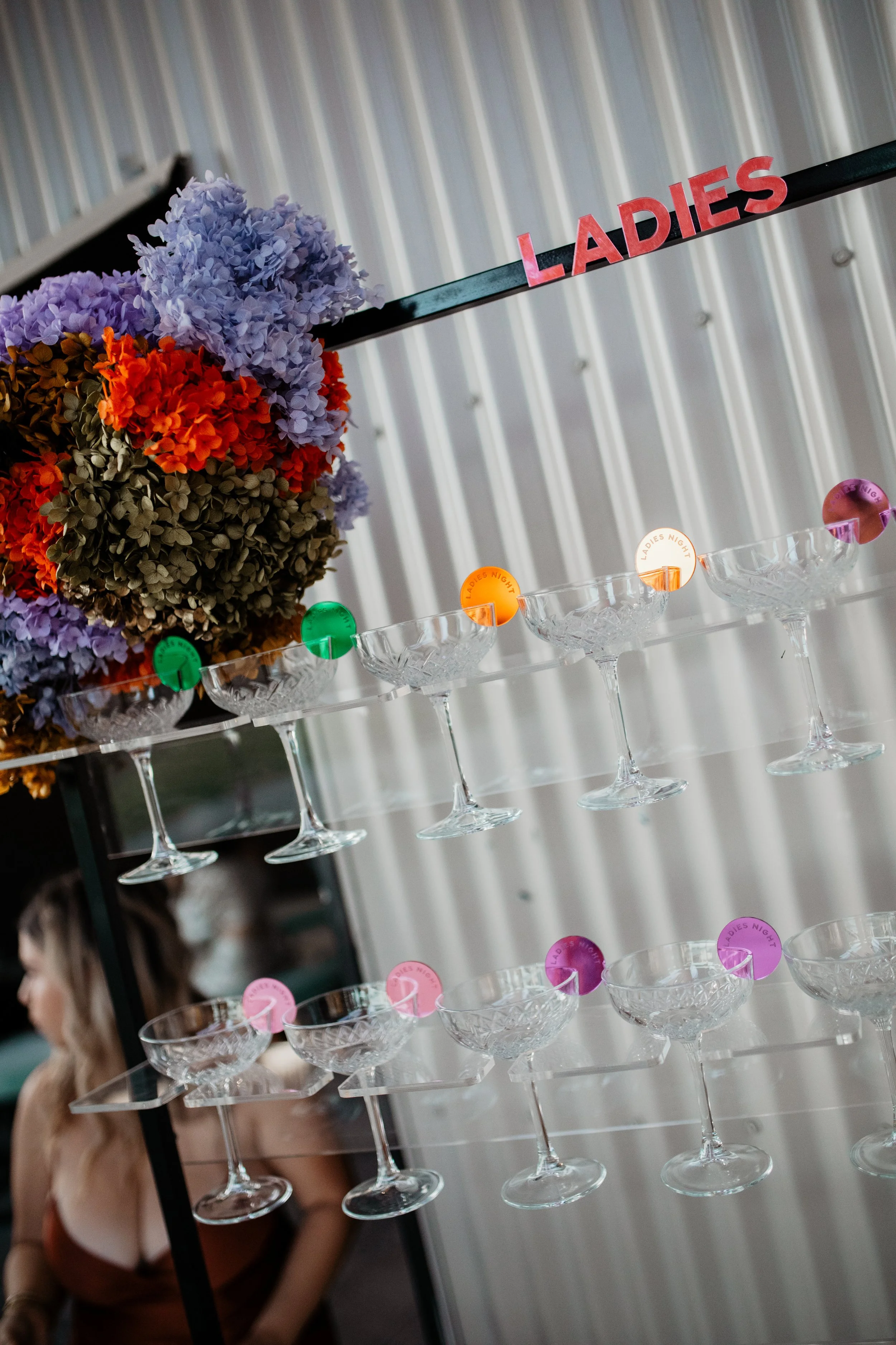 A display with glassware and colorful labels for a ladies' night event, decorated with purple, red, and orange flowers.
