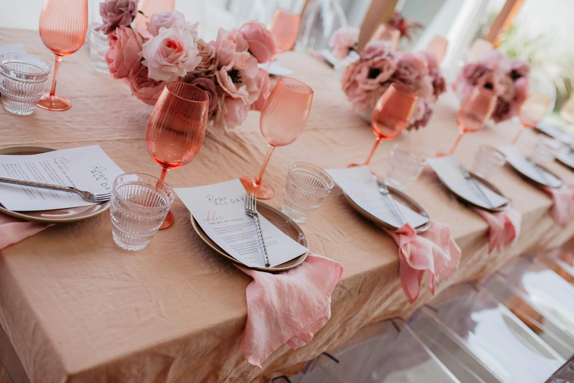 Elegant dining table decorated with pink flowers, pink glasses, and gold-rimmed plates and utensils, set for a special occasion in natural light.