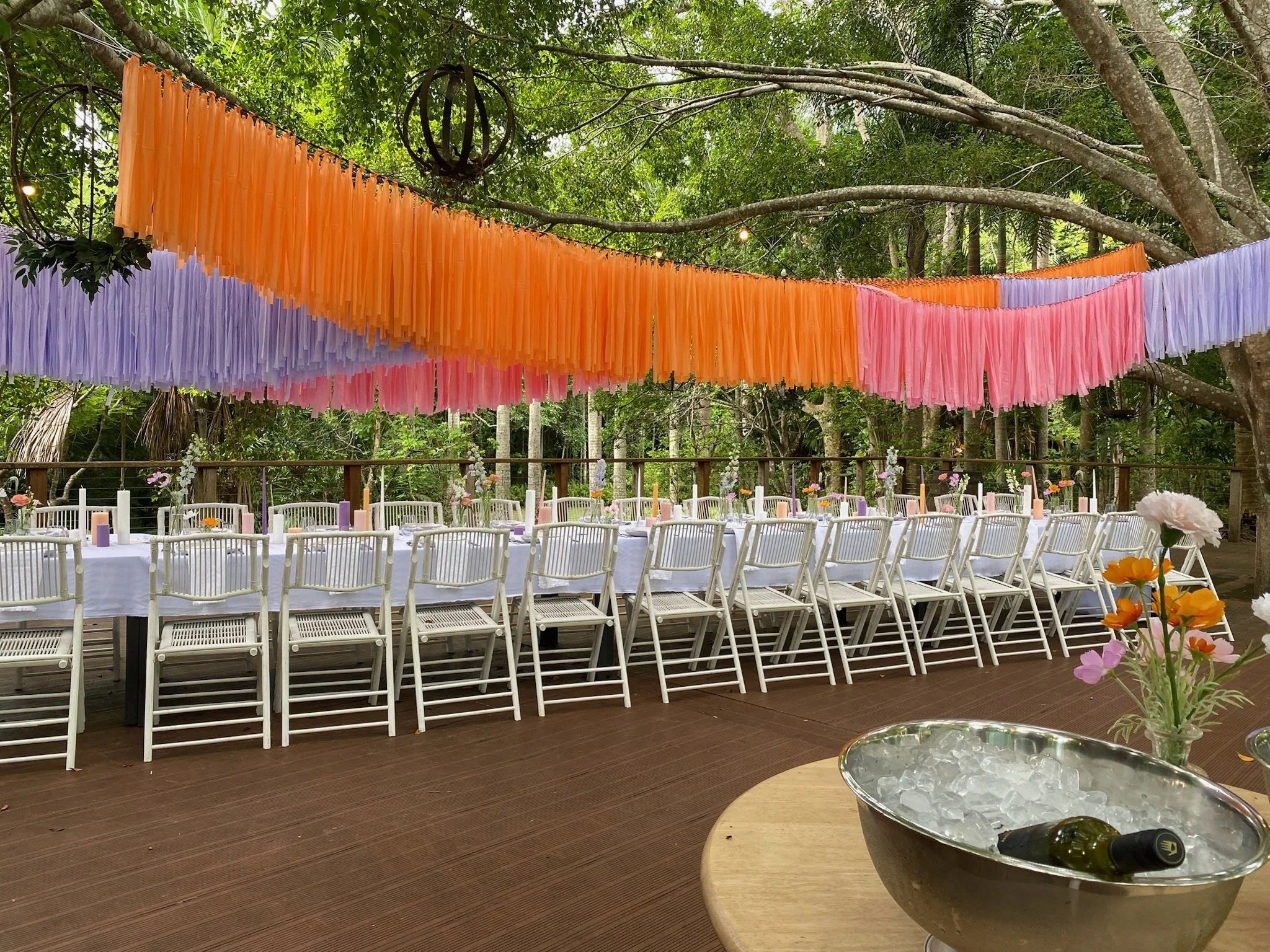 Outdoor event setup with long white tables and white chairs, decorated with pastel-colored flowers and candles, under hanging orange, pink, and purple streamers, surrounded by trees in a forest setting, with a metal ice bucket and bottle in the foreg