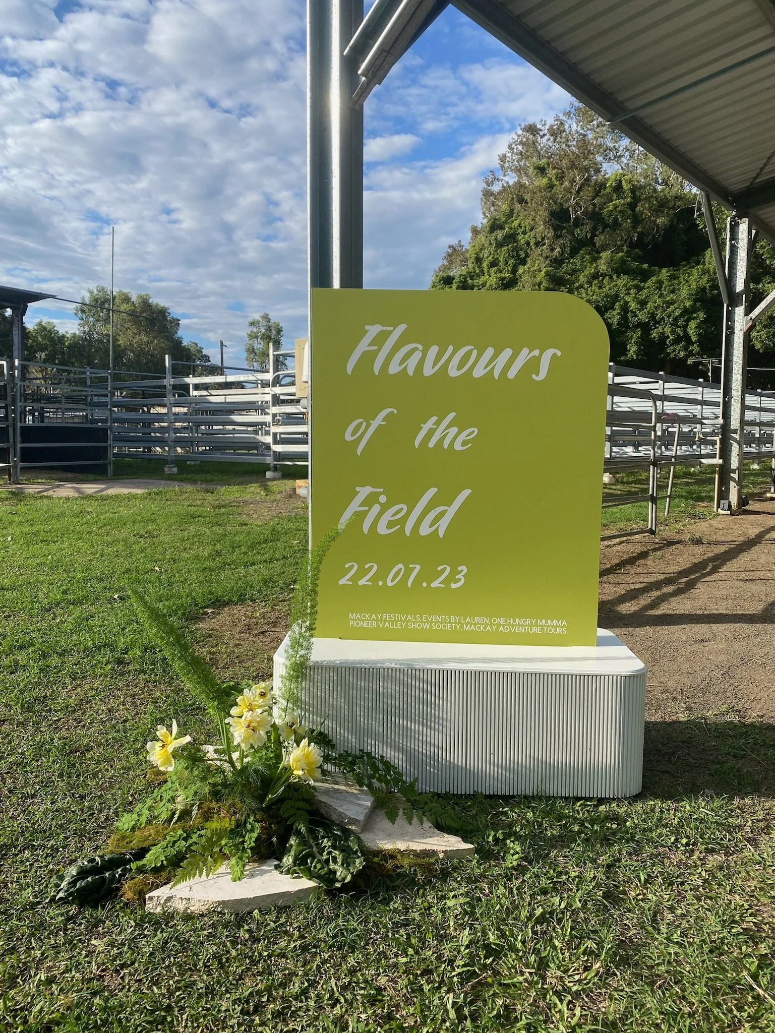 A large, yellow-green sign reading 'Flavours of the Field 22.07.23' is placed on a grassy area near a sports field with metal bleachers, a canopy, and a blue sky with clouds in the background. There are decorative flowers and greenery at the base of 