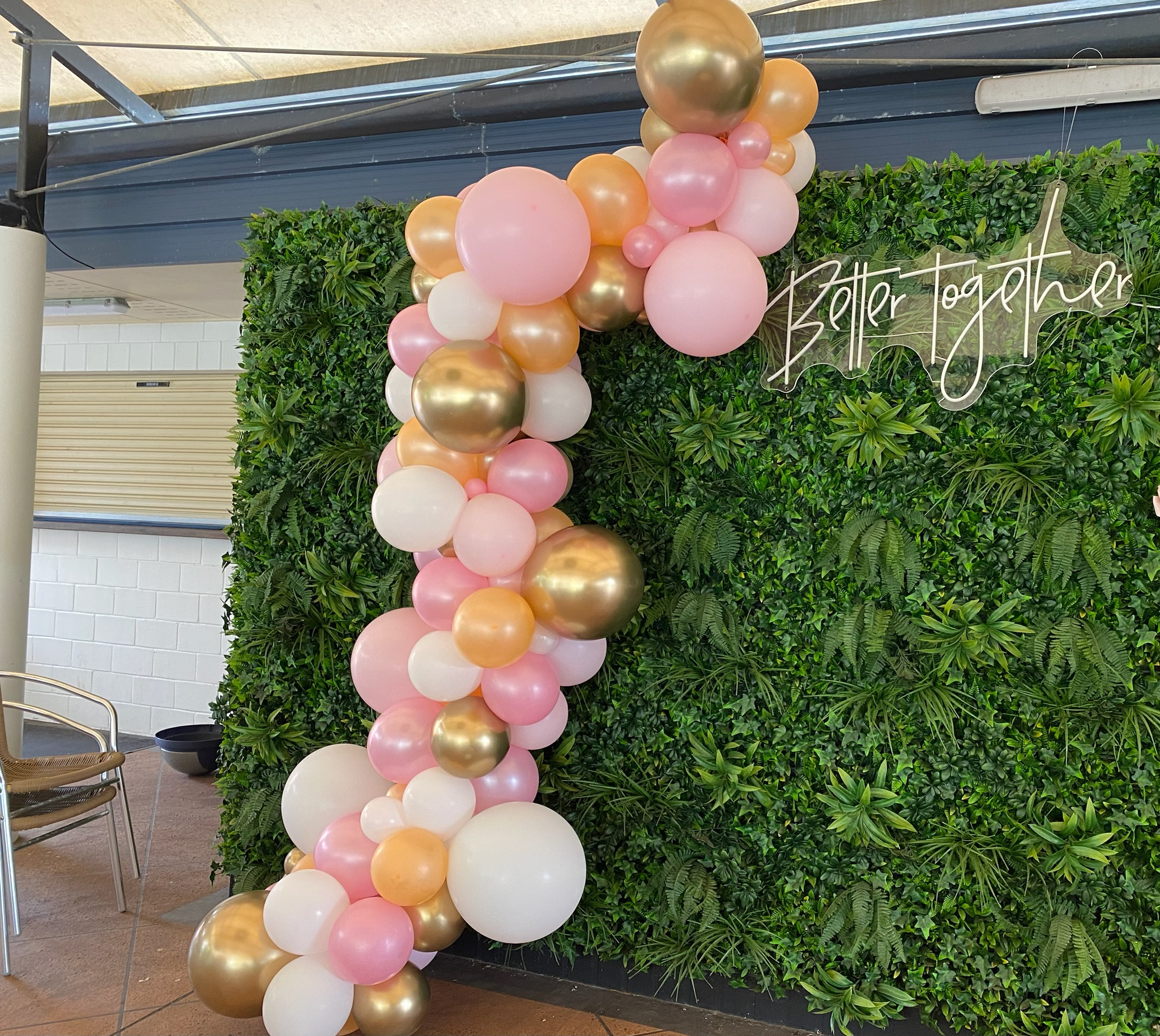 Decorative display of pink, white, gold, and peach balloons arranged as a garland in front of a green leafy wall with a neon sign that says 'Better Together'.