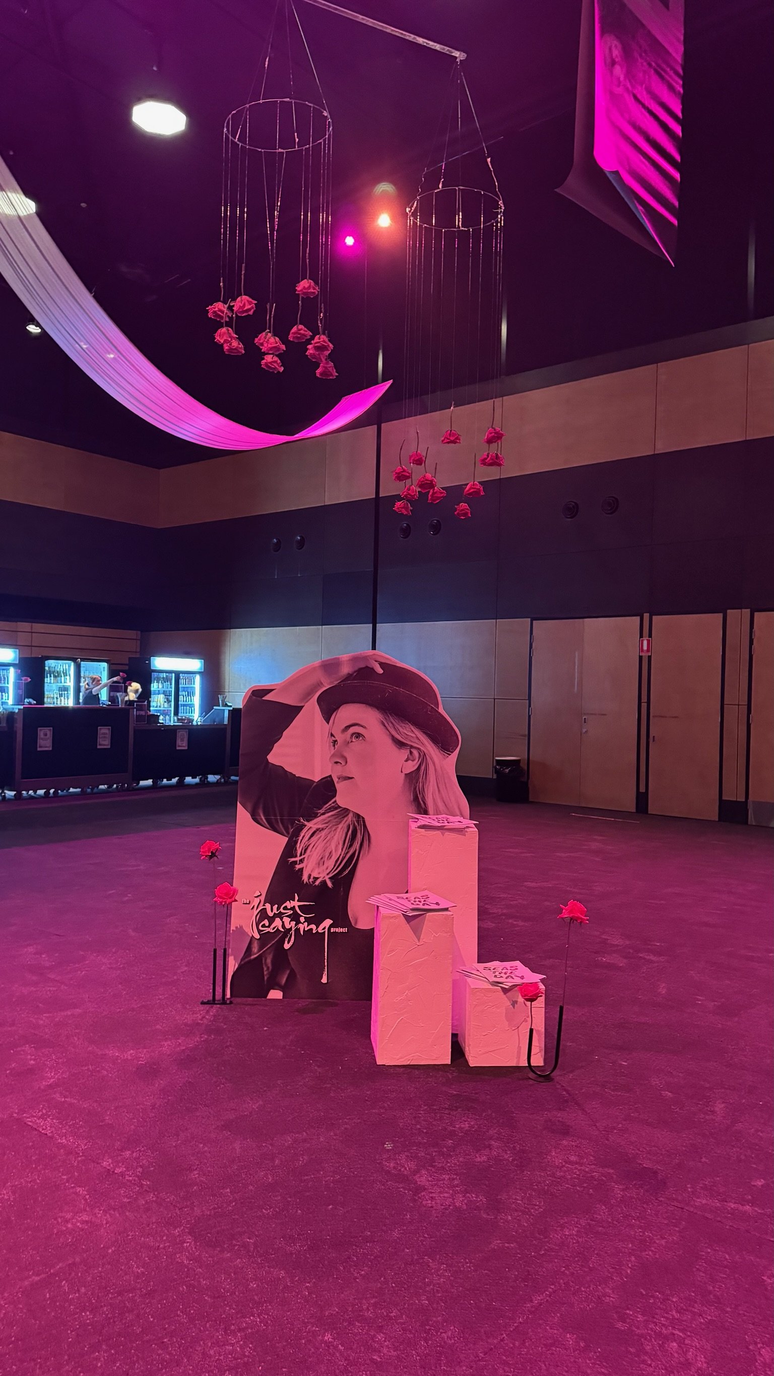 An indoor event space decorated with pink lighting, hanging pink flowers, and a large black-and-white poster of a woman wearing a hat, with the text "Just Saying" on it.