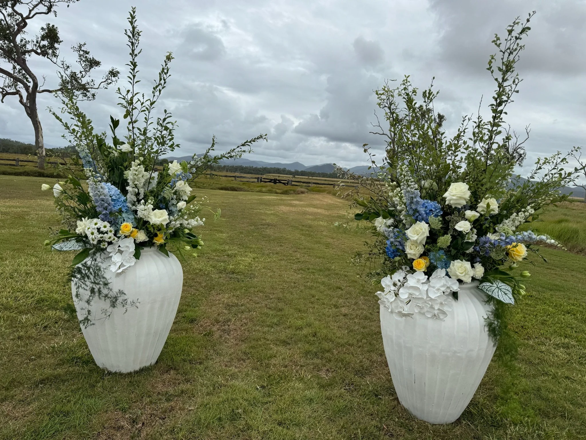 Two large white vases filled with white, blue, and yellow flowers are placed on grass outdoors on a cloudy day, with a tree, wooden fence, and mountains in the background.