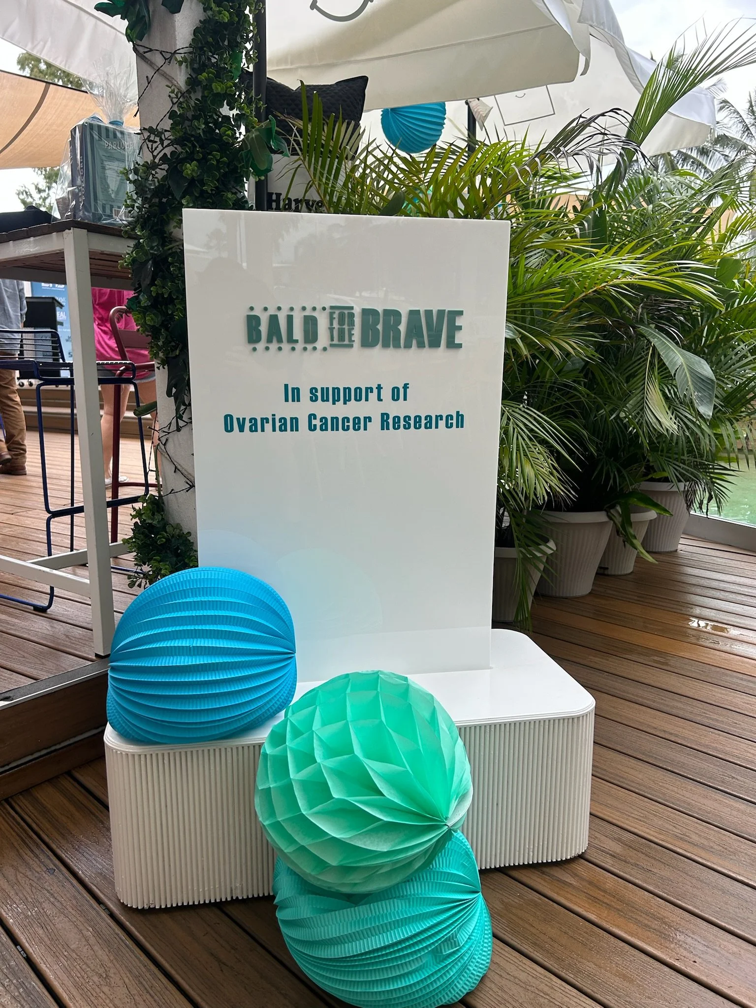 Sign supporting 'Bald for the Brave' campaign for ovarian cancer research, decorated with blue and green paper lanterns, placed on a white bench at an outdoor event with plants and umbrellas in the background.
