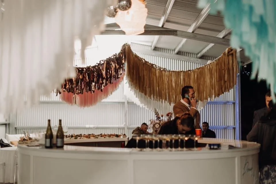 Indoor event with decorations made of hanging paper and fabric fringe streamers, a man on a cellphone, bottles of wine or champagne on a counter, and guests seated in a decorated venue.