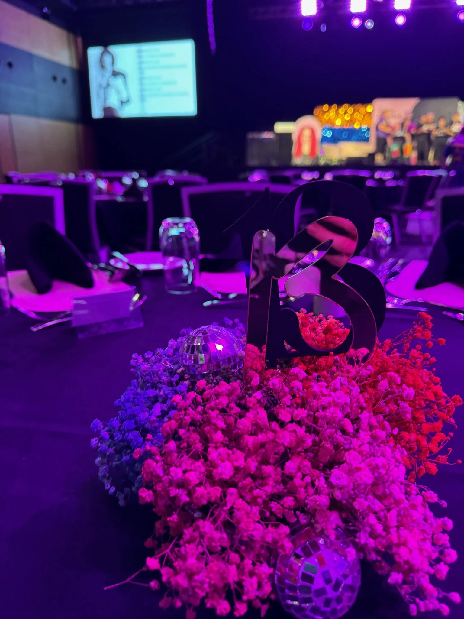 Table centerpiece with pink flowers and large black number 3 at an event or celebration, with a blurred background of a stage and seating area.
