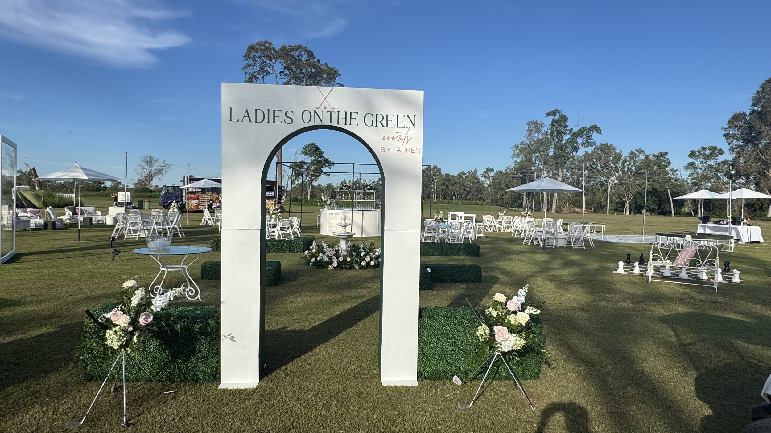 Outdoor event setup with white chairs, umbrellas, and floral arrangements on a lawn with trees in the background. There is a sign reading "Ladies on the Green" and a white arch with pink flowers.