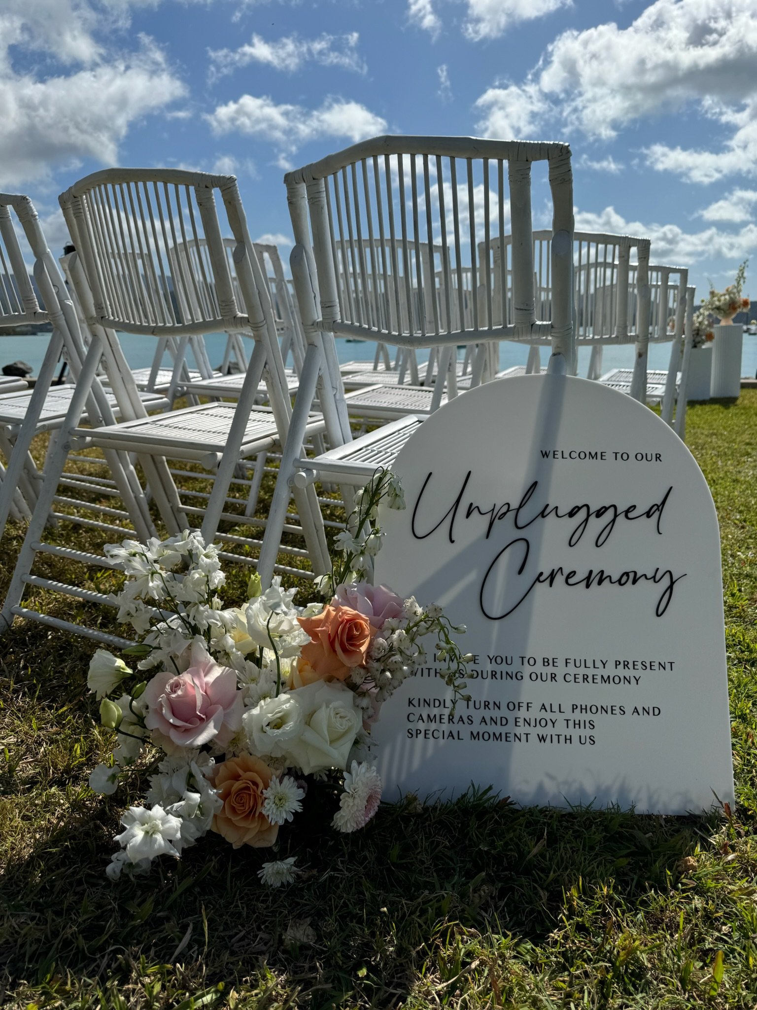 Outdoor wedding ceremony setup with white chairs arranged on grass near a body of water, a floral arrangement of pink and white roses and daisies, and a white sign with black cursive text reading 'Unplugged Ceremony' on a sunny day with clouds in the