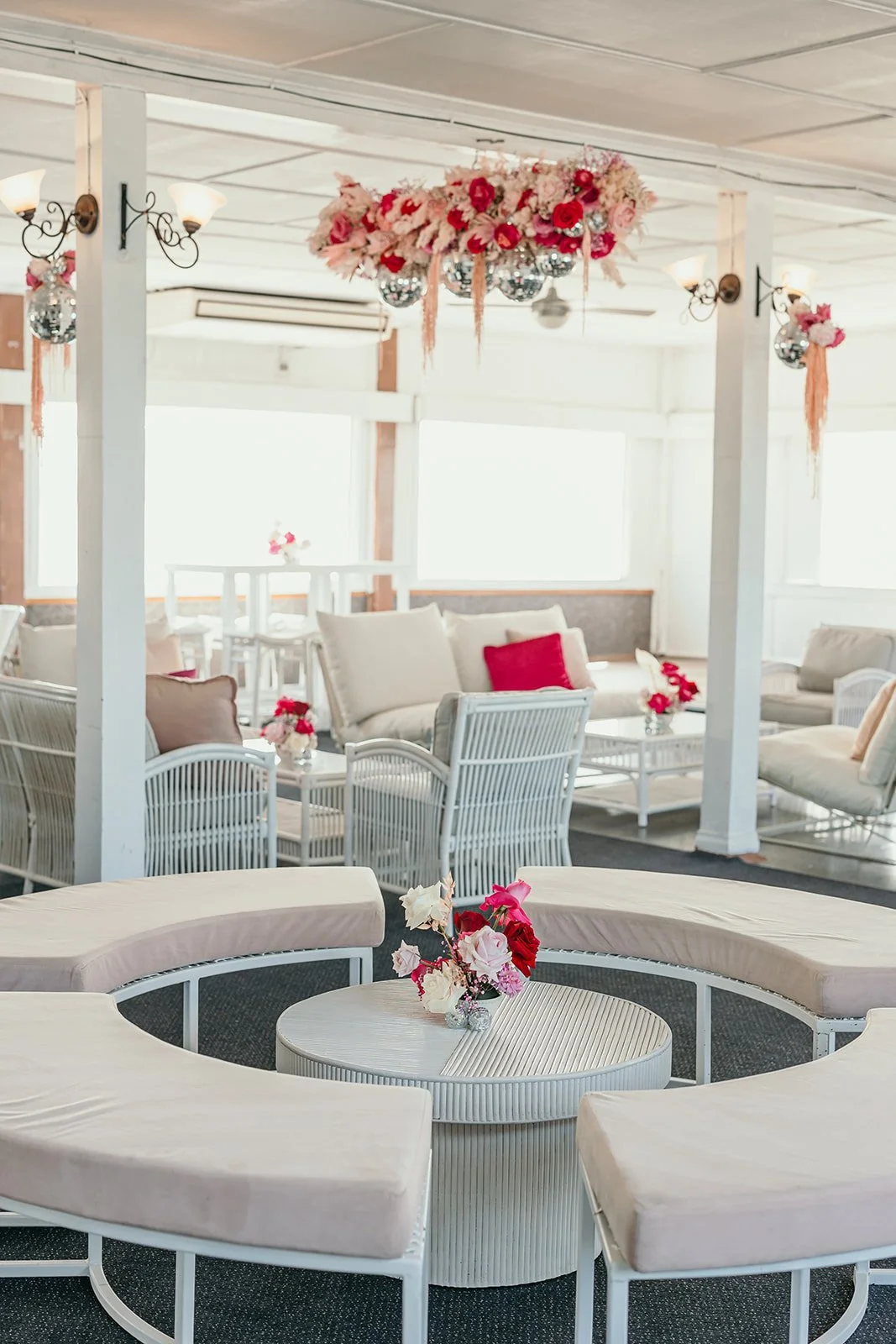 Indoor lounge area decorated with pink and red flowers, including a floral chandelier and small flower arrangements on tables, with white furniture and large windows.