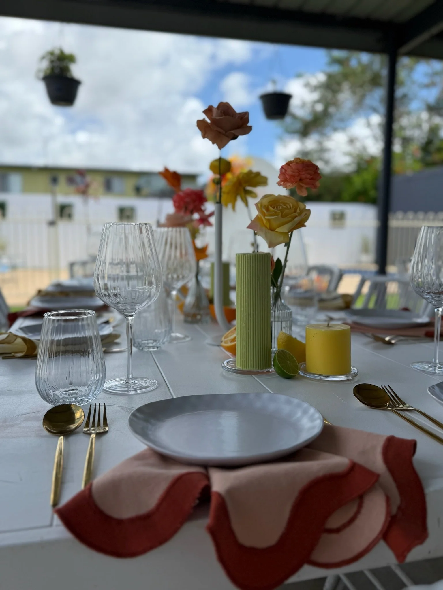 Decorated outdoor dining table set with wine glasses, plates, cutlery, colorful flowers, candles, and napkins under a covered patio.