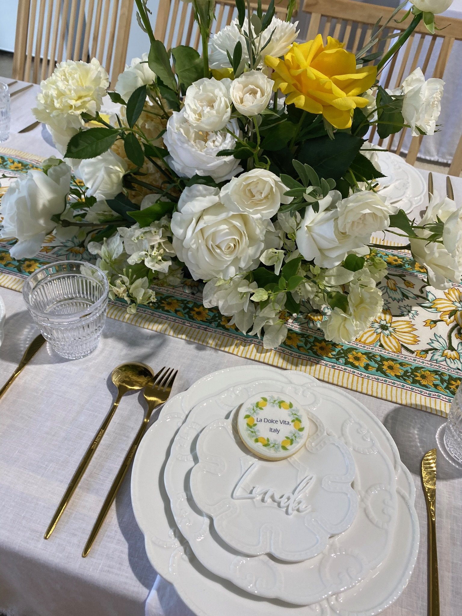 A table setting with a floral centerpiece of white and yellow flowers, gold utensils, clear glasses, and a decorative white plate with a lemon-themed cookie and the word 'Lunch' on it.