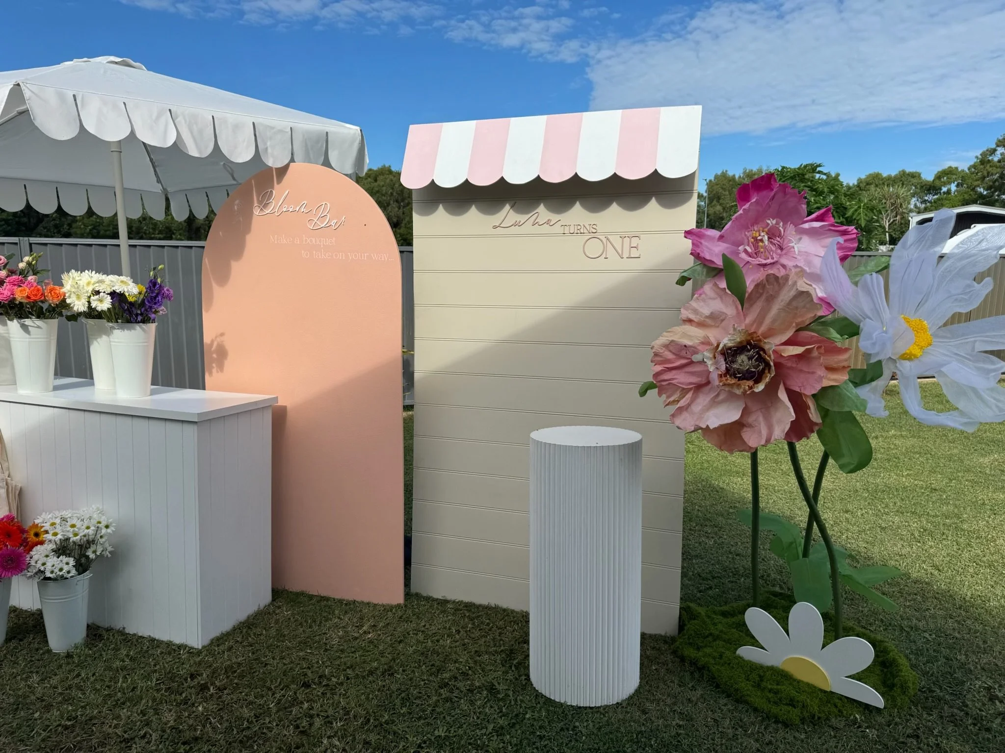 Decorative outdoor setup with pastel-colored boards, flower arrangements, a striped canopy, and a large artificial flower arrangement for a child's first birthday celebration.
