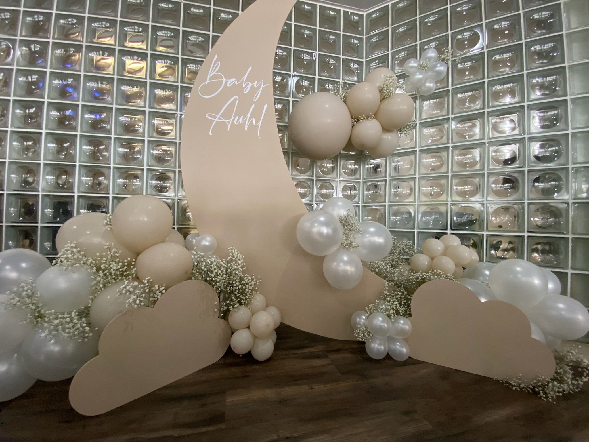 Decorative baby shower setup with white and beige balloons, white flowers, and a beige moon-shaped backdrop with 'Baby Auhl' written on it, against a glass block wall.
