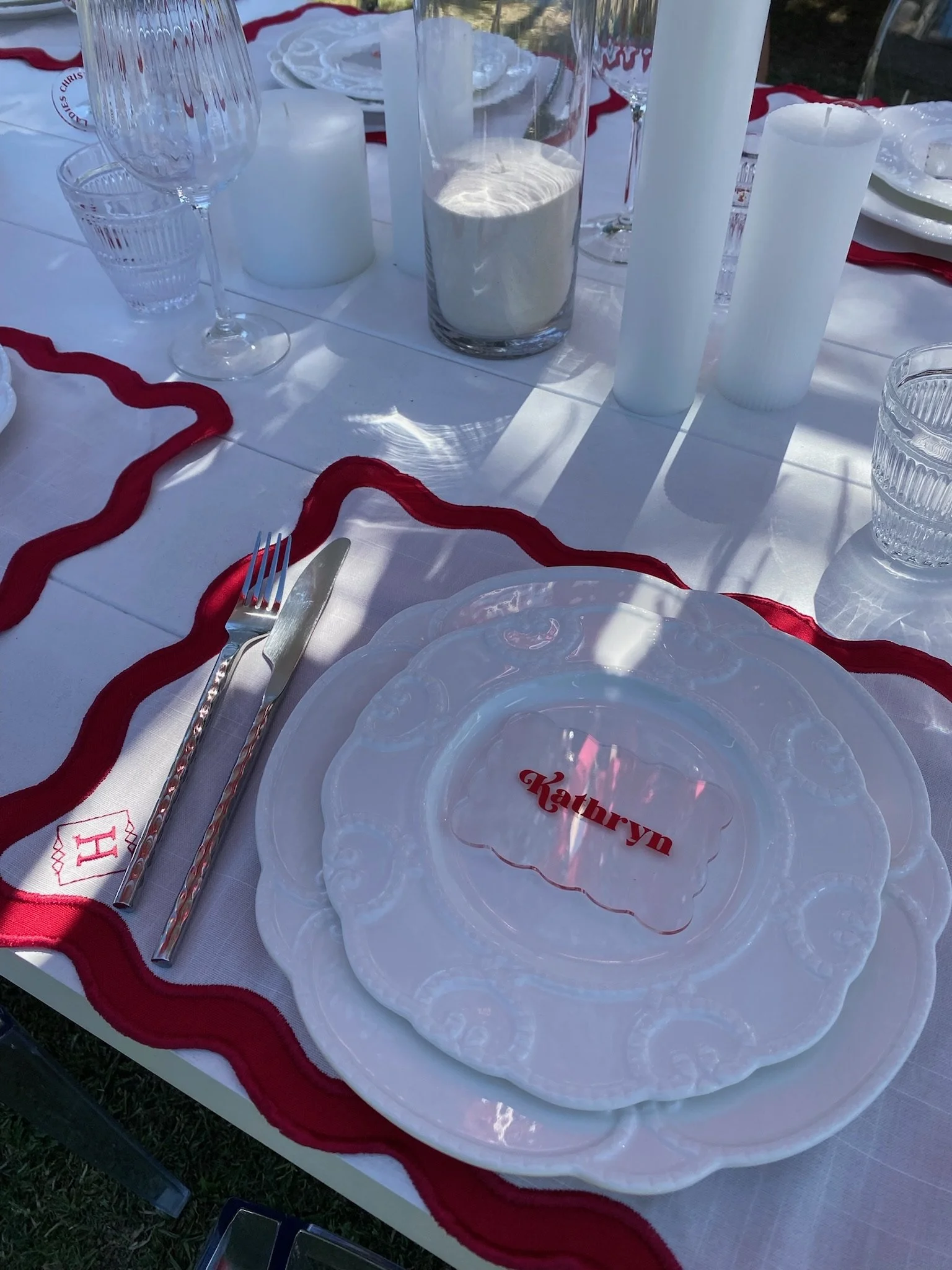 A table setting with white plates, silverware, a wine glass, candles, and a place card with the name 'Kathryn' on it, decorated with red accents.
