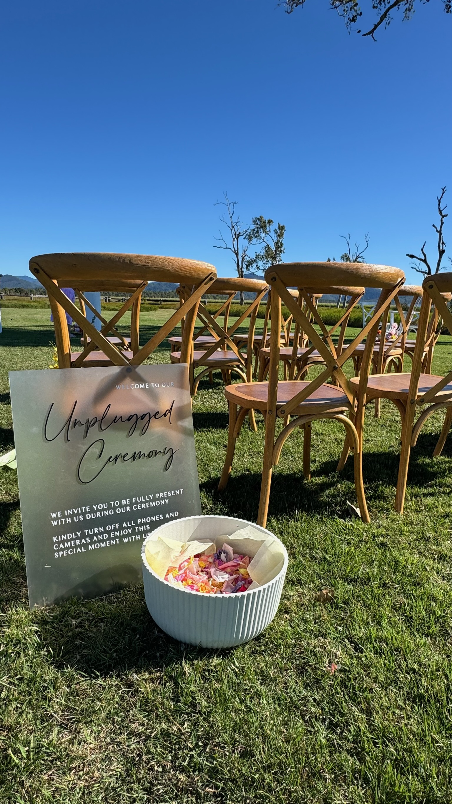 Outdoor wedding ceremony setup with wooden chairs arranged on grass, a welcome sign that reads 'Unplugged Ceremony', and a bowl of flower petals, under a clear blue sky.