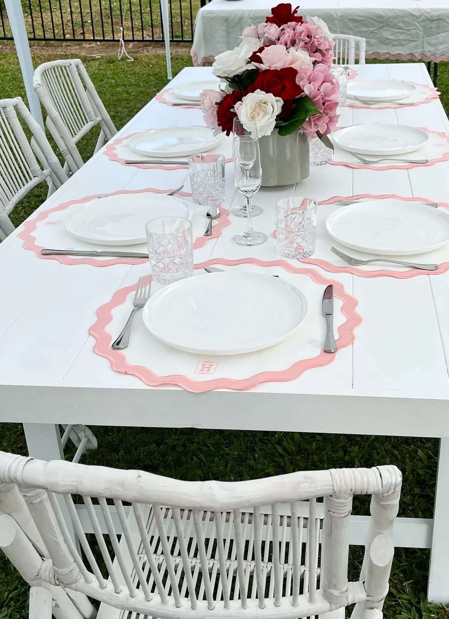 White outdoor dining table set for a meal with pink and white floral centerpiece, pink scalloped placemats, white plates, silverware, and crystal glasses, surrounded by white chairs on a grassy lawn.