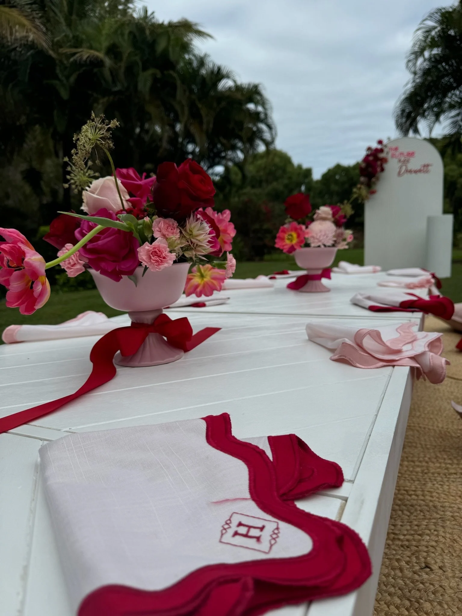 A decorated outdoor table with pink and red flower arrangements in pink vases, set with folded napkins and ribbons, suggesting a celebration or wedding event.