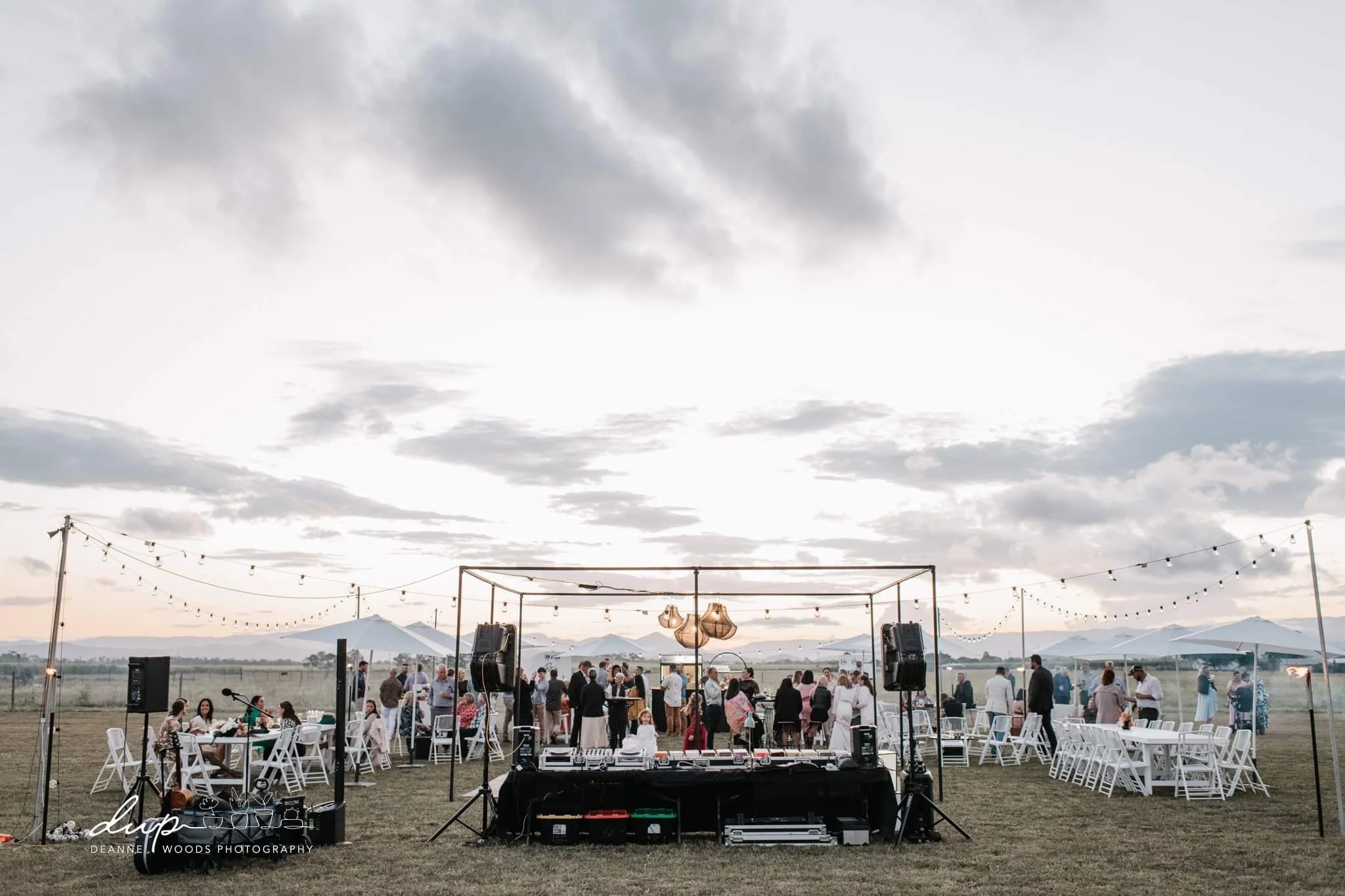 Outdoor wedding reception setup with guests, white tables and chairs, string lights, a DJ booth, and an open field with mountains in the background during sunset.