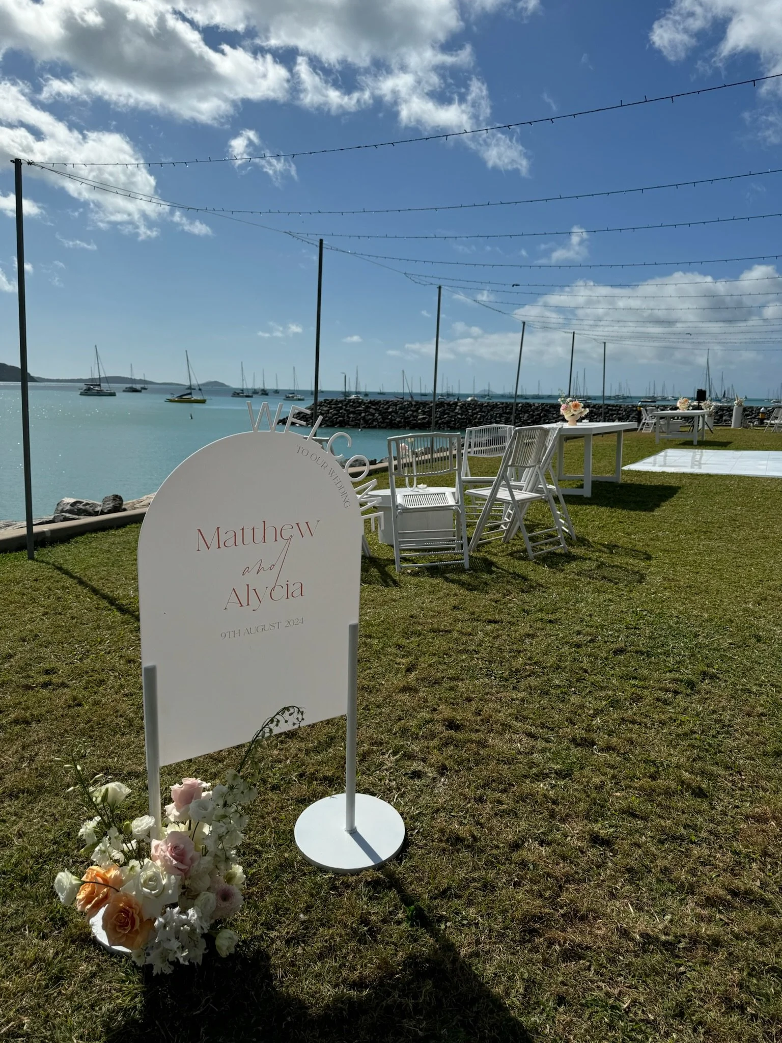 Wedding setup on a grass lawn near a body of water with sailboats, white chairs, and a sign reading 'WELCOME TO OUR WEDDING Matthew W and Alycia 9th August 2024' with a flower arrangement nearby.