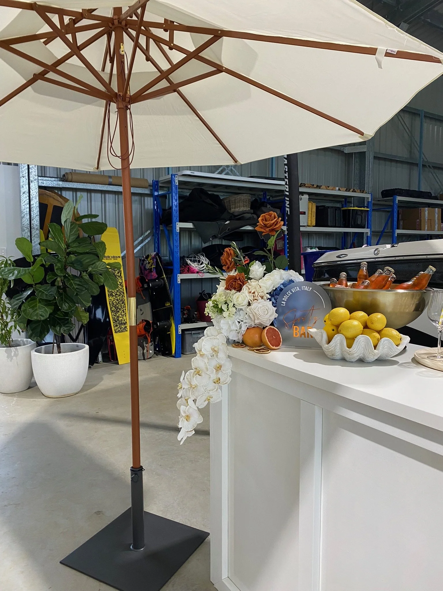 Outdoor setup with large white umbrella, potted plant, bar area with flowers, citrus fruits, and bottled drinks.