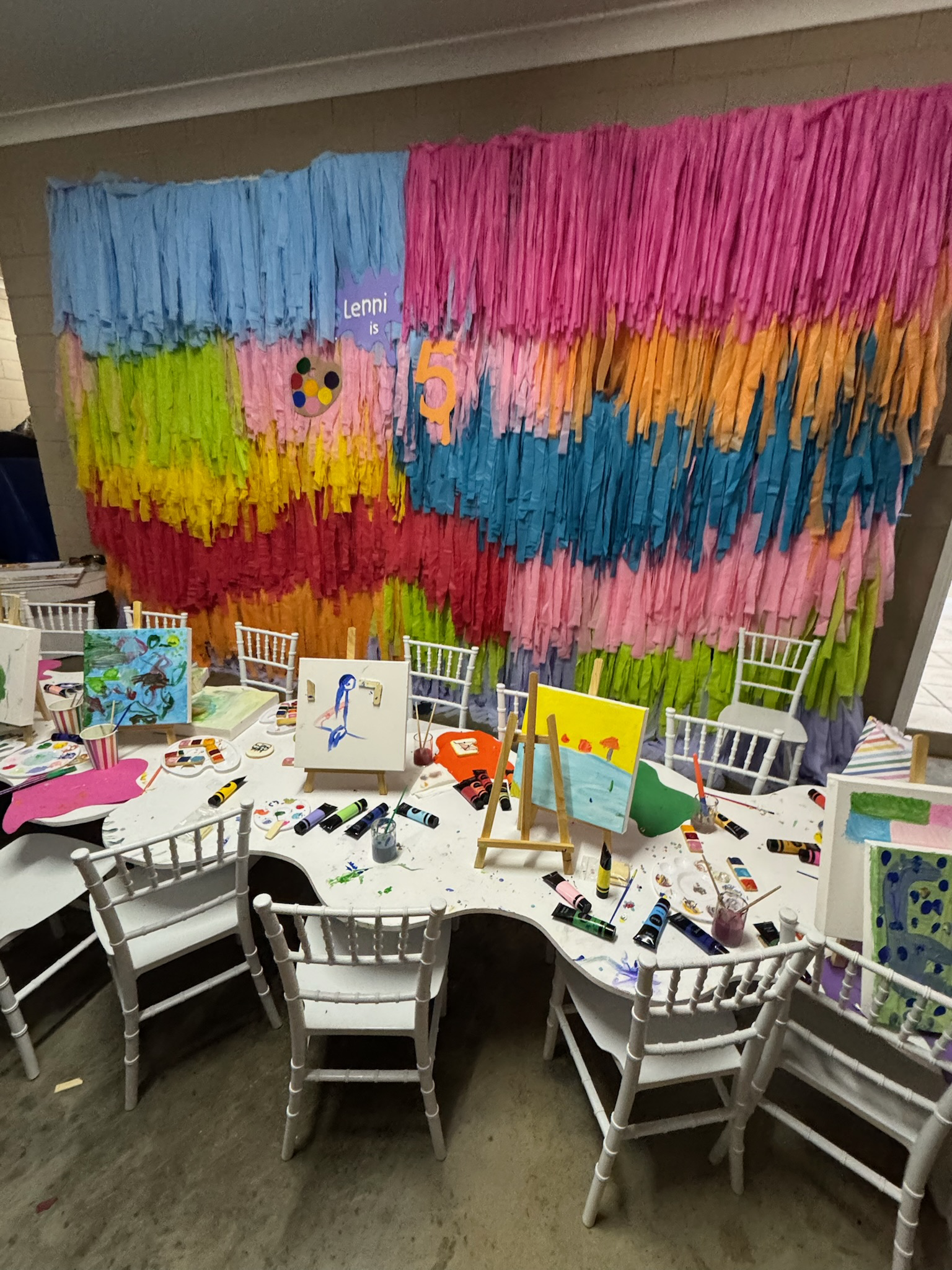 Decorated party room with a colorful rainbow paper backdrop, a table with painting supplies and completed art projects, and white chairs around the table.