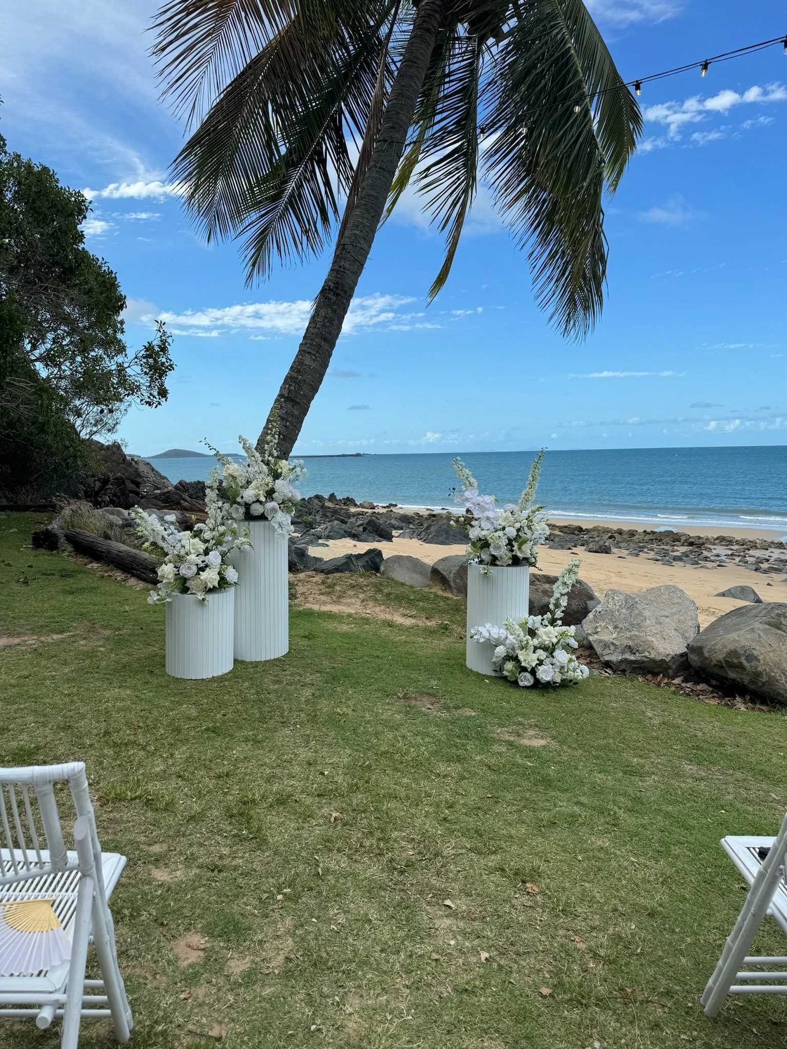 A beachside outdoor wedding setup with white floral arrangements in tall white vases, white chairs, a leaning palm tree, and a view of the ocean and rocks under a blue sky.