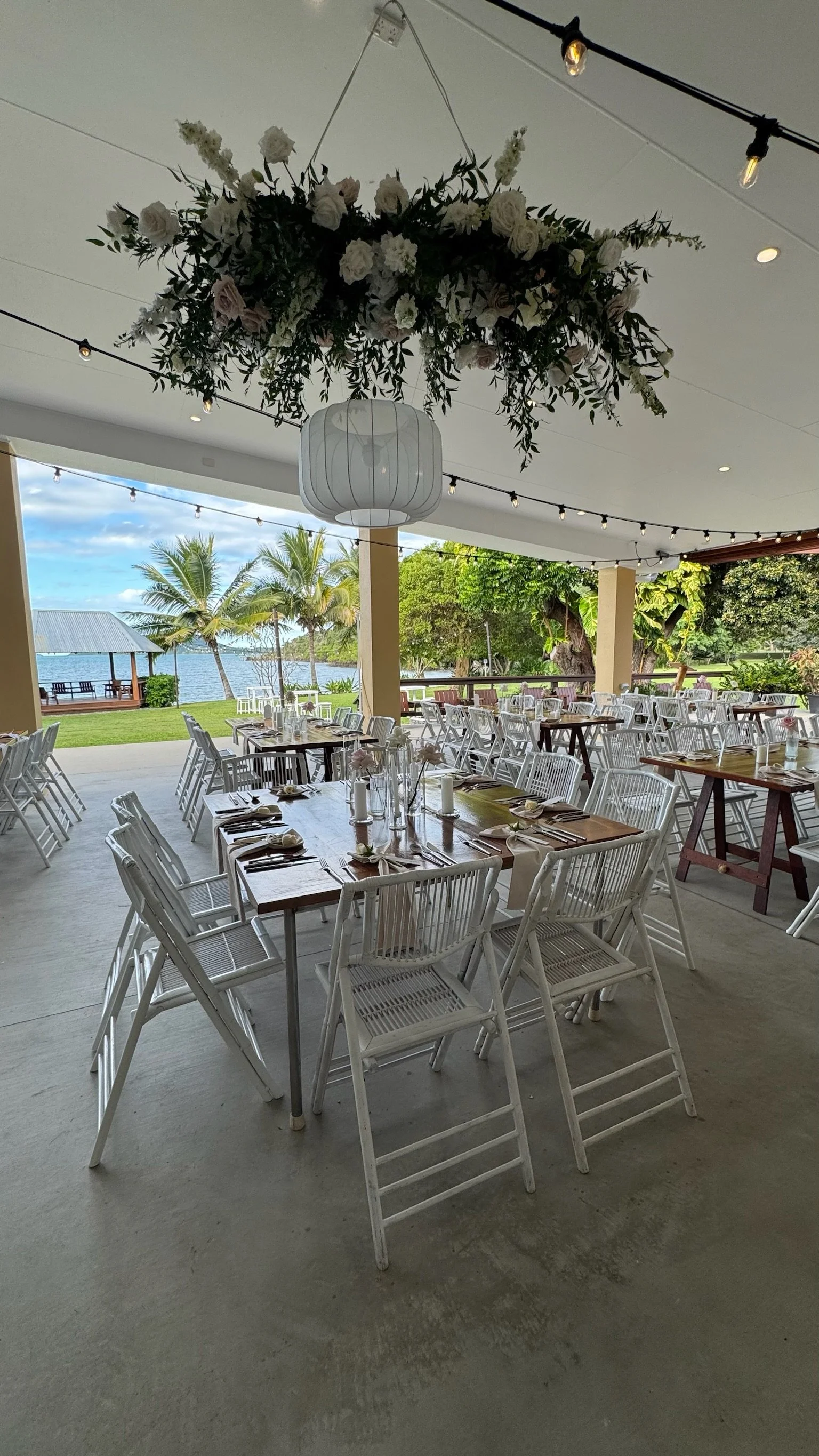 Wedding or event reception setup with tables and chairs set near a waterfront, featuring decorative hanging floral arrangement and string lights.