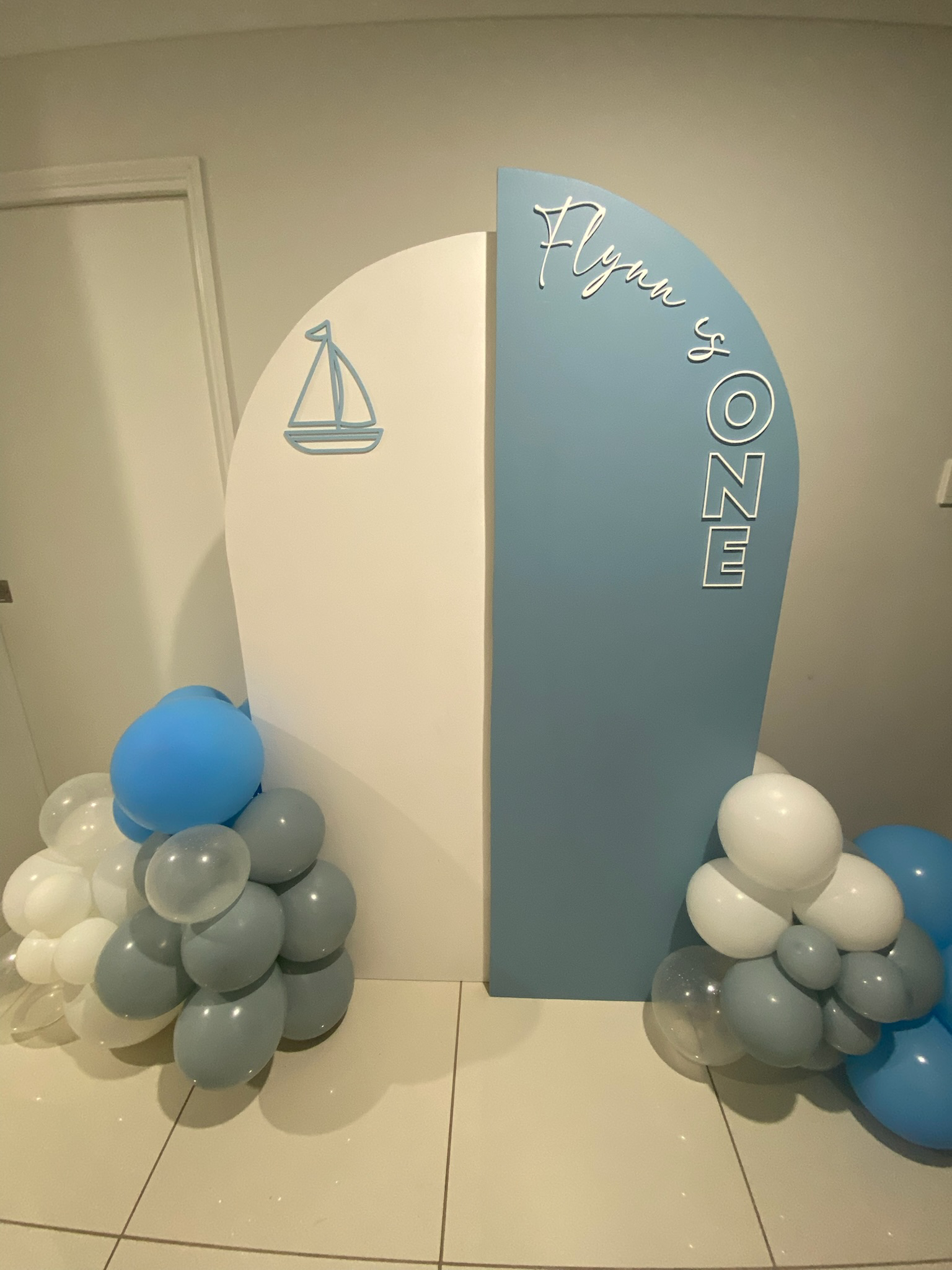 Decorative celebration backdrop with blue and white panels, a sailboat icon, and balloons in shades of blue, white, and gray.