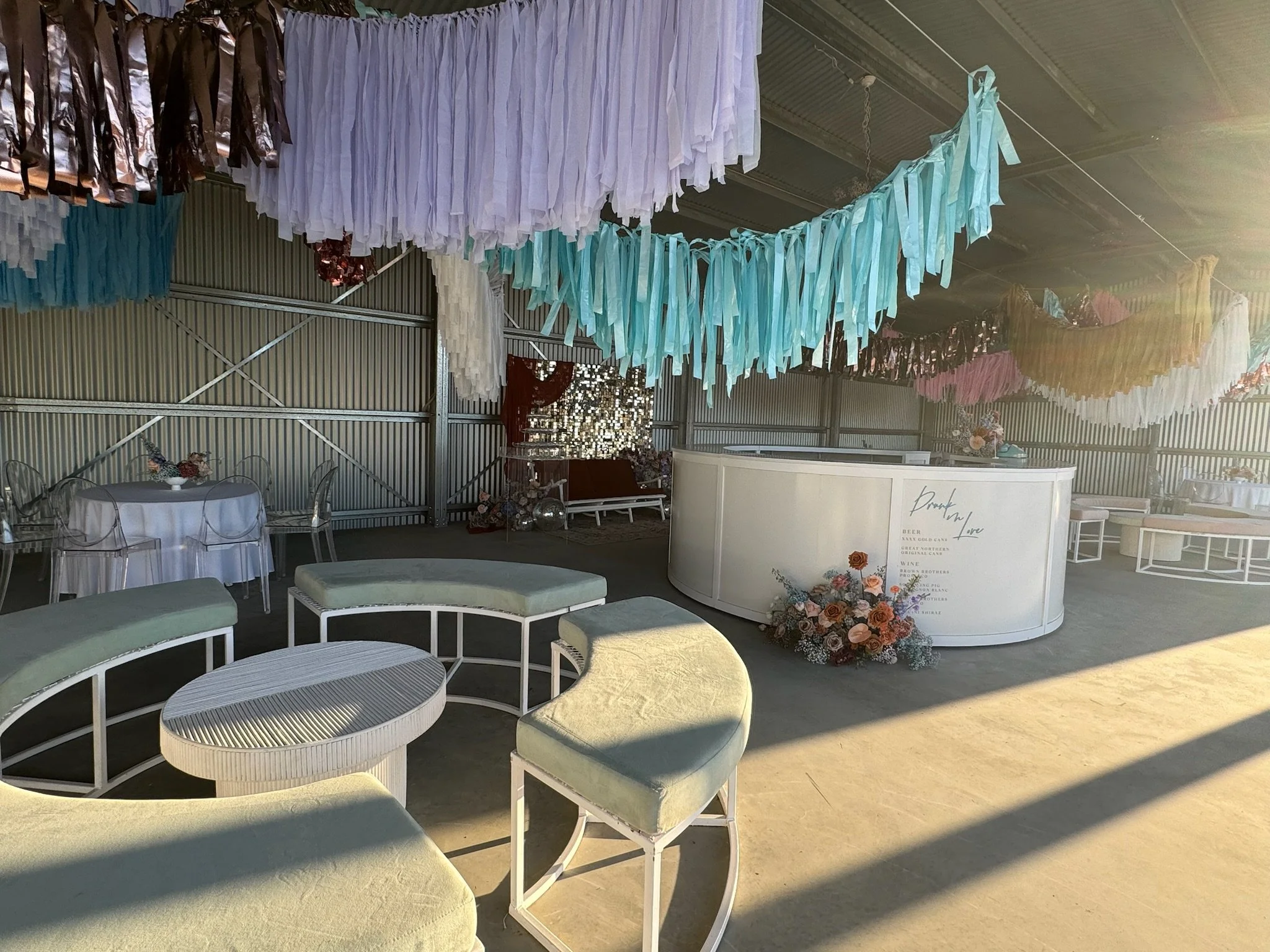 Decorated event space with pastel-colored tassel garlands hanging from the ceiling, white circular bar with floral arrangement, and seating area with curved couches and tables, sunlight illuminating the space.