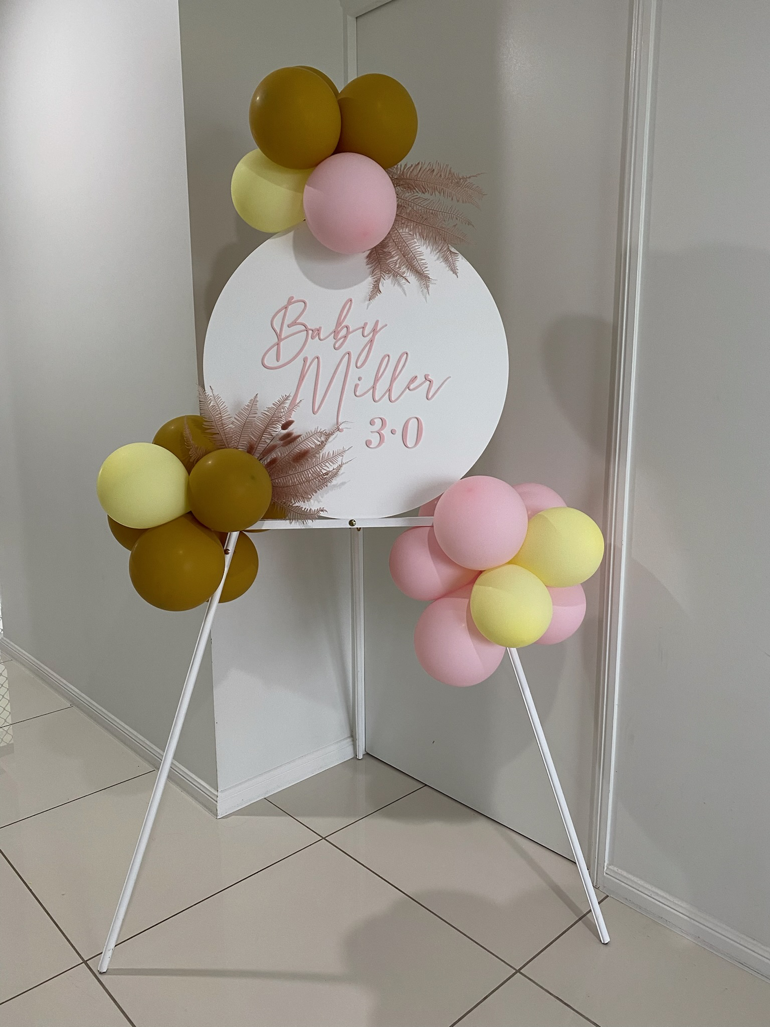 Decorative baby announcement sign with pink and yellow balloons, dried pink leaves, and white tripod stand, announcing Baby Miller 3.0.