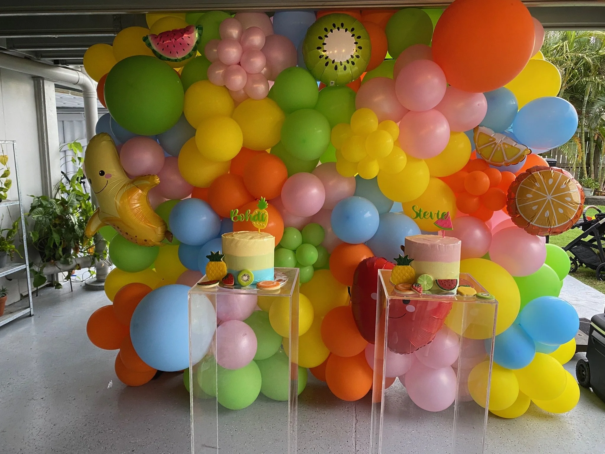 Colorful balloon backdrop with various shaped balloons, including fruit-themed balloons, two cakes with tropical fruit toppers on clear stands, and themed decorations with labels 'Bohdi' and 'Stevie.'