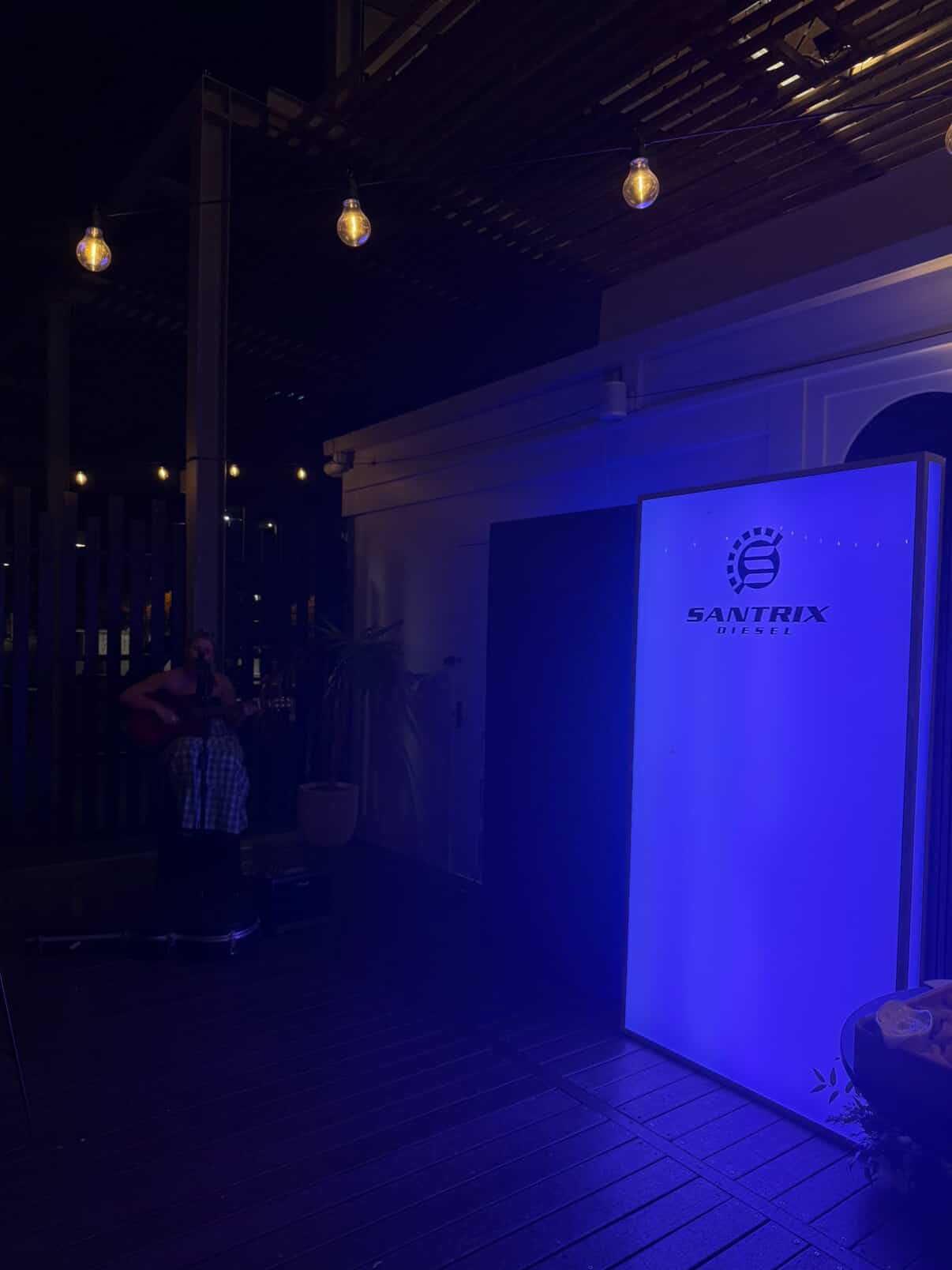 Nighttime outdoor scene with string lights hanging from a wooden pergola. A person with a guitar, dressed in checkered shorts, is standing on the left. A large illuminated sign displays the logo and name 'Saltrix Diesel' on the right.