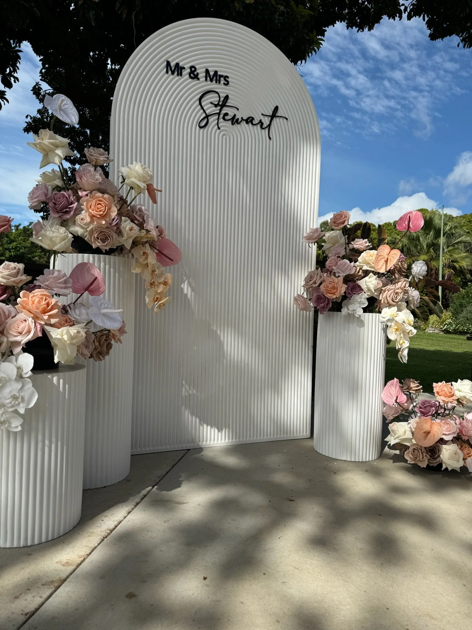 Wedding backdrop with white ribbed panels, floral arrangements of pink, white, and purple roses, and greenery, with a sign reading 'Mr & Mrs Stewart' in black cursive. Outside on a sunny day with a blue sky and trees in the background.