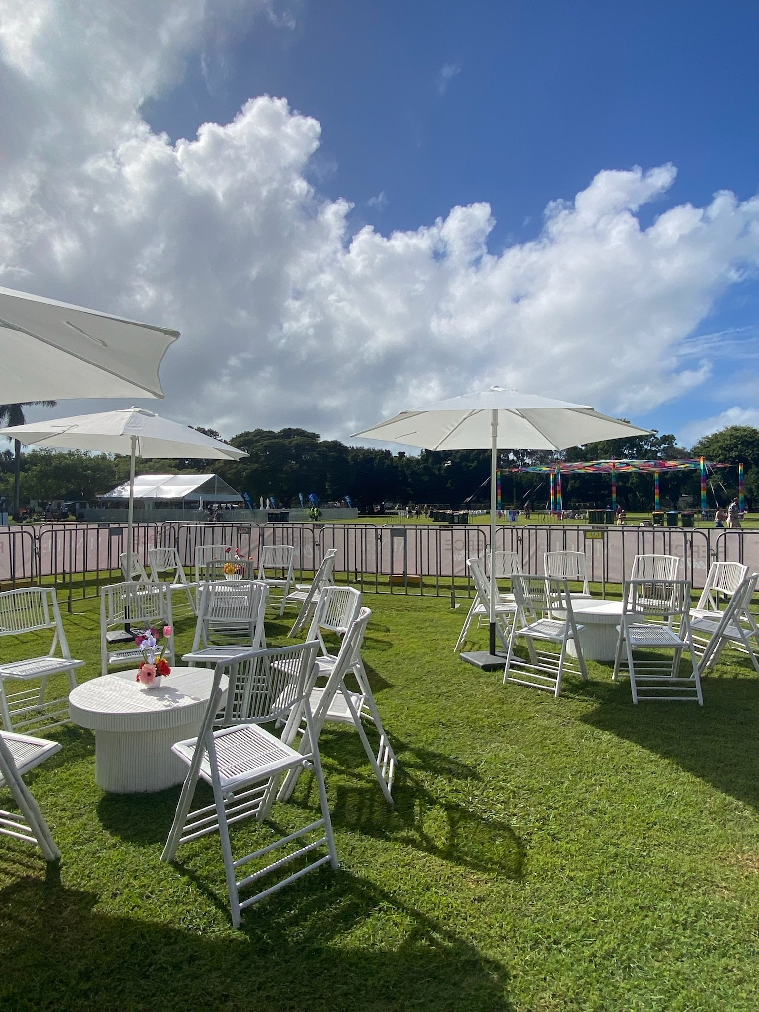 Outdoor event setup with white tables, chairs, and umbrellas on green grass, with a fairground and cloudy sky in the background.