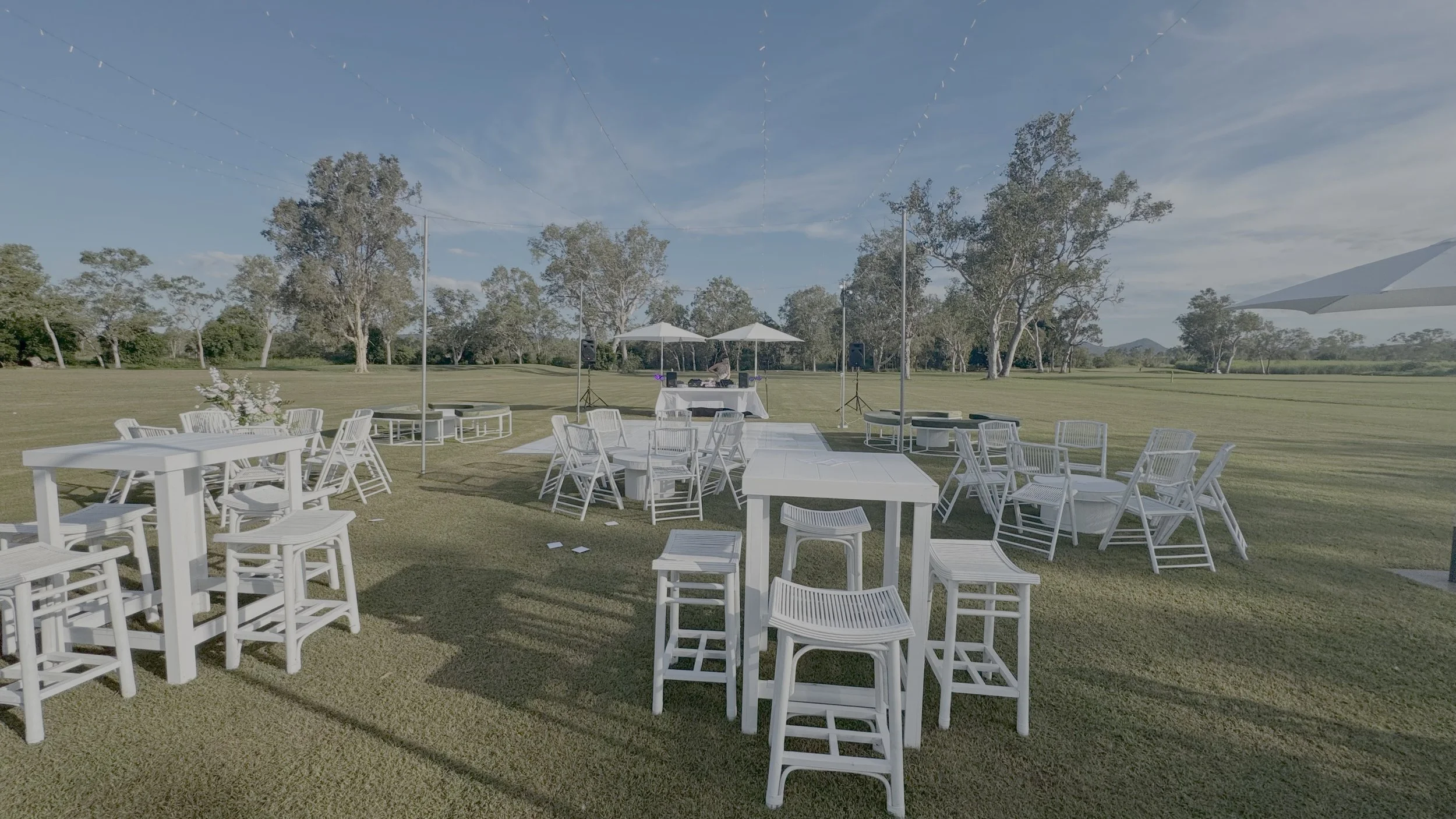 Outdoor event setup on a grassy field with white tables and chairs, two white umbrellas, and a DJ booth with speakers in the background, surrounded by trees and a blue sky.