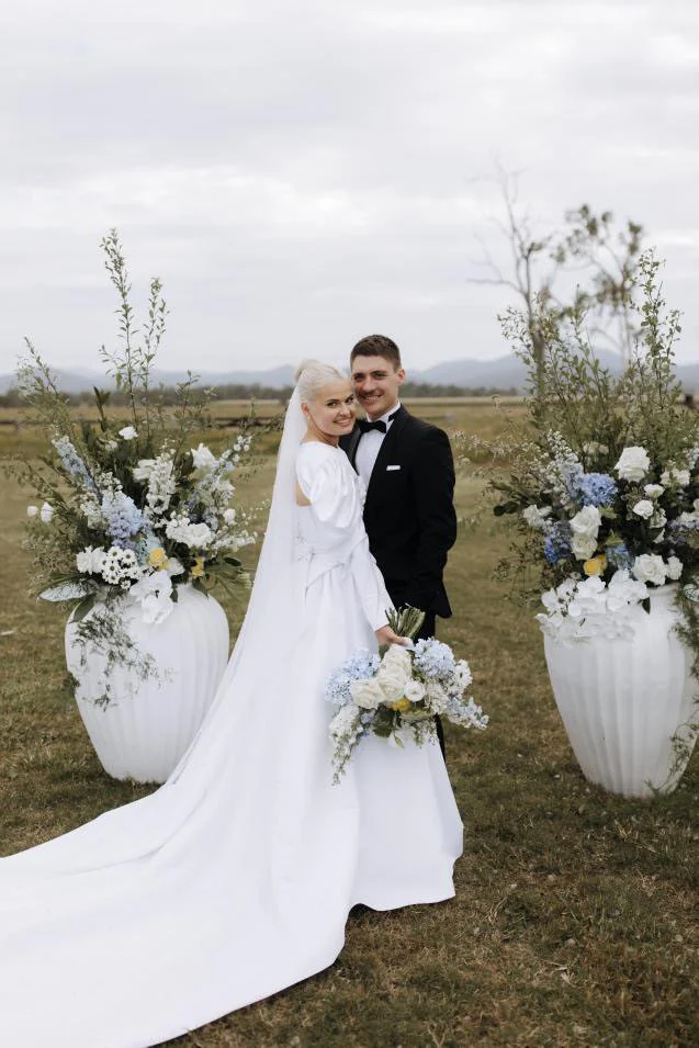 Bride and groom standing together outdoors with large floral arrangements in white vases on either side, holding a bouquet, in a countryside setting.