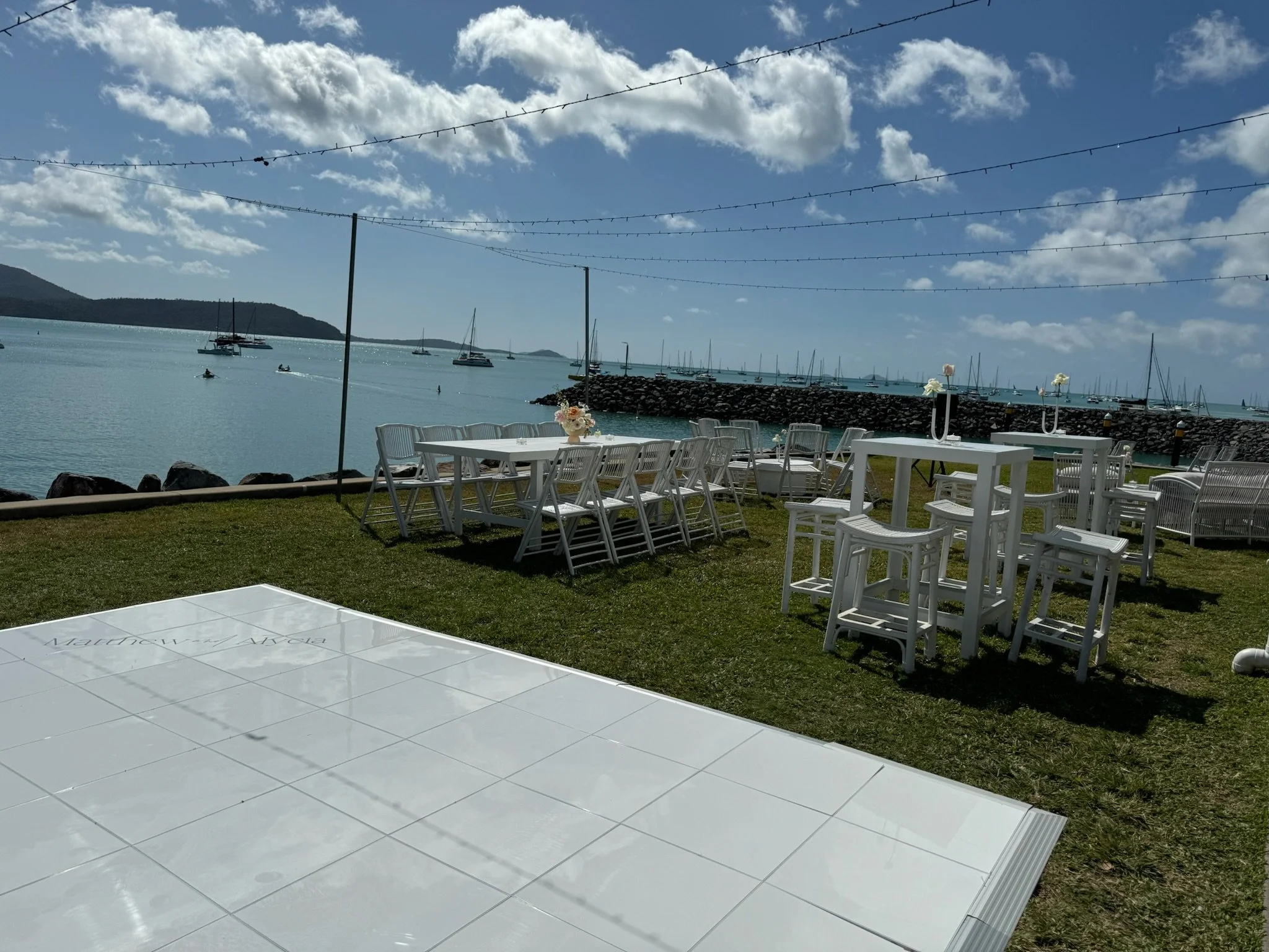 Outdoor patio area with white chairs and tables overlooking a marina with sailboats, on a partly cloudy day.