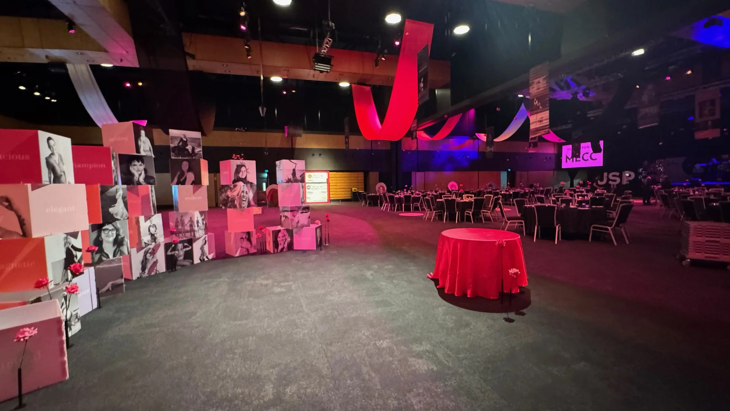 Event space decorated with pink and red theme, with tables and chairs, large display of boxes with photos of women, and banners with text including 'JSP', 'MECC', and 'champion'.