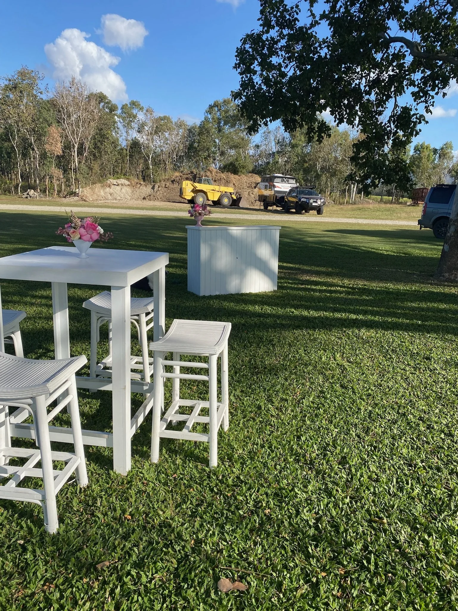 Outdoor setting with white high tables and chairs, decorated with pink floral arrangements, on a grassy area under a tree with a construction site and trees in the background.