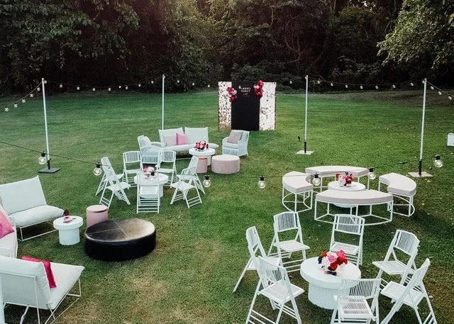 Outdoor event setup with white furniture, string lights, and floral decorations on tables, arranged on a grassy field surrounded by trees.